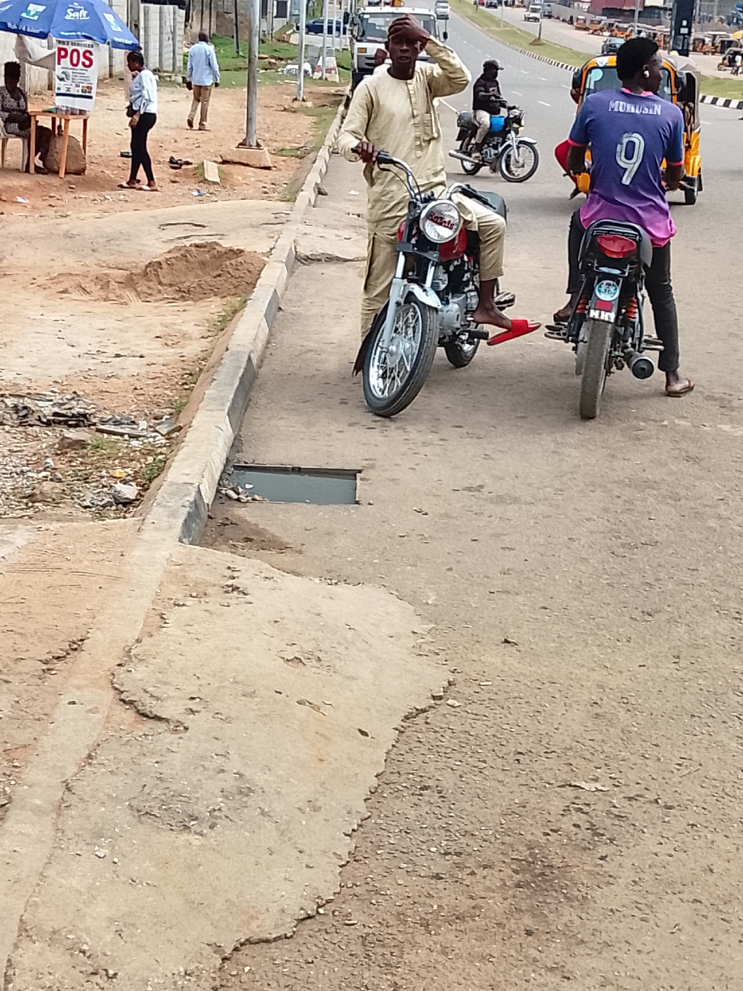 The image depicts a street scene in what appears to be a lower-income area, potentially on the outskirts of Abuja, Nigeria. Two men are on motorcycles, one in a purple jersey with the name "Murusin" and the number 9 on the back, and the other in a traditional tan outfit. One man is adjusting his cap. Several other people and vehicles, including motorcycles, auto rickshaws, and cars are visible in the background. A "POS" (Point of Sale) kiosk is present on the side of the road, suggesting commercial activity. The road is worn, and the surrounding area is sandy and unpaved. A drainage opening can be observed. The overall environment suggests a bustling but somewhat informal urban setting.