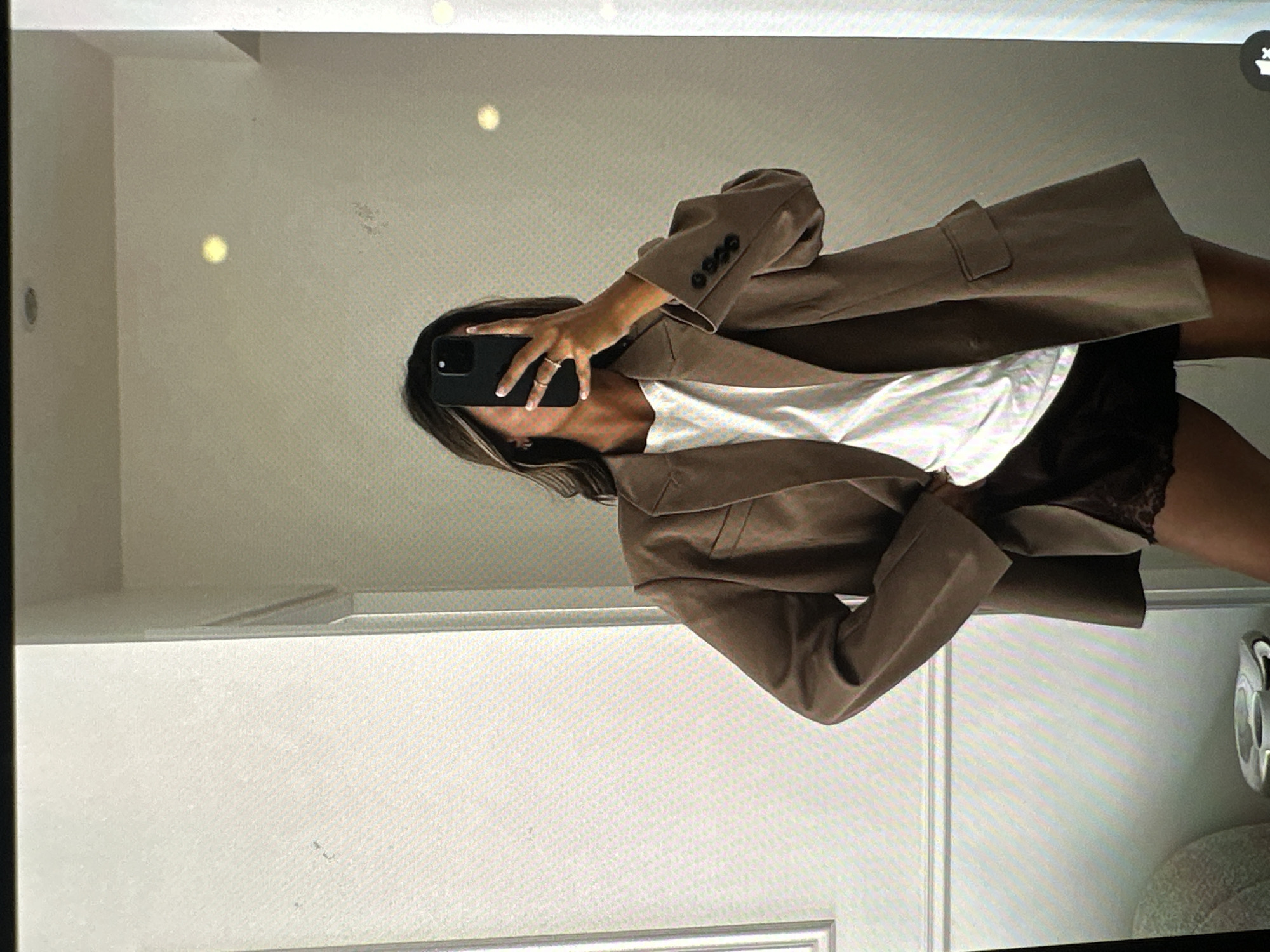 A person is taking a mirror selfie. The person has long, dark hair with blonde highlights and is wearing a brown blazer over a white t-shirt and black shorts. They are holding a black smartphone in their right hand, obscuring their face. The location appears to be a room with white walls, a white door, and a glimpse of what appears to be a light fixture or a sink. There is no information present to suggest this photo was taken in Yerevan, Armenia.