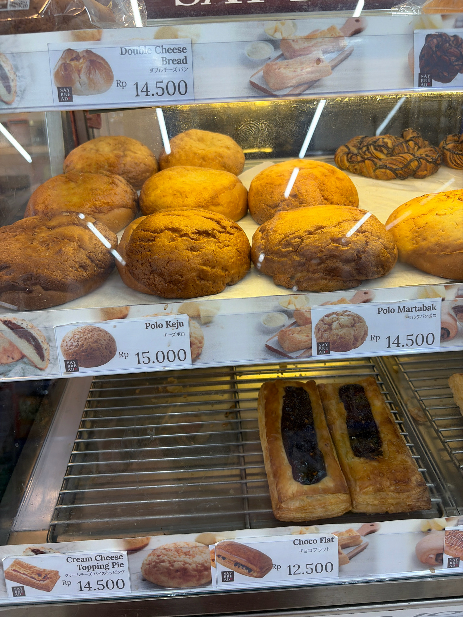 The image displays a bakery display case filled with various types of bread and pastries. The display is well-lit, with fluorescent lights illuminating the products. In the foreground, on the bottom shelf, are two rectangular pastries with a dark filling, labeled "Choco Flat" with a price of Rp 12,500. To the left, there's a pastry labeled "Cream Cheese Topping Pie" priced at Rp 14,500, and to its right, a round pastry.
Above this shelf, on the middle shelf, are several round, golden-brown buns. A label below these reads "Polo Keju" (Cheese Polo) with a price of Rp 15,000 and also "Polo Martabak" (Martabak Polo) priced at Rp 14,500, with accompanying images of these items.
On the top shelf, a sign indicates "Double Cheese Bread" with a price of Rp 14,500, showing an image of a cheese-filled bread. Other baked goods are visible in the background, including braided pastries with chocolate drizzle and other buns.
The overall environment appears to be a commercial bakery, likely in Tanjung Pinang, Indonesia, given the Indonesian Rupiah (Rp) currency used for pricing. The display case is clean and organized, showcasing a variety of bakery items.