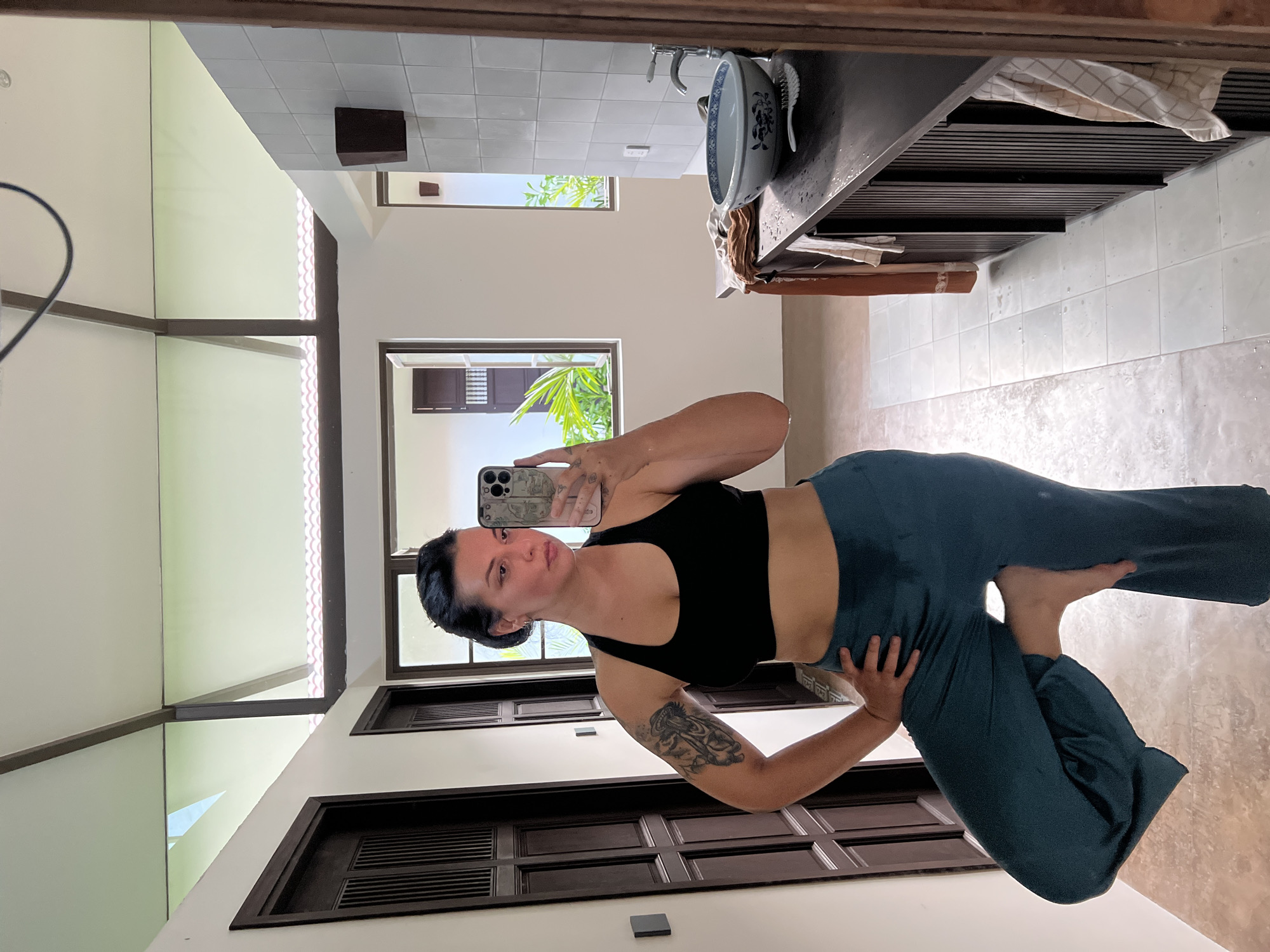 This is a portrait-oriented, vertically oriented, medium shot photograph of a woman taking a mirror selfie in what appears to be a bathroom or dressing area. The woman has dark hair pulled back, is wearing a black sports bra and teal colored pants, and has tattoos on her left arm. She is standing with her left hand on her hip and her right leg raised, balancing on her right foot. She is looking at her phone, which she is holding in her right hand, and her expression is neutral.
The background of the image is a reflection in a mirror. The reflection shows a tiled floor, white walls, and dark wooden doors. There are windows visible in the reflection, one of which shows lush green foliage, suggesting an outdoor tropical environment. A sink with a blue and white patterned bowl and a faucet is visible on a dark countertop, along with a towel and a brush. The overall impression is of a bright, well-lit space, possibly in a hotel or resort, given the tropical greenery and clean aesthetic. The time of day appears to be daytime due to the natural light coming through the windows.