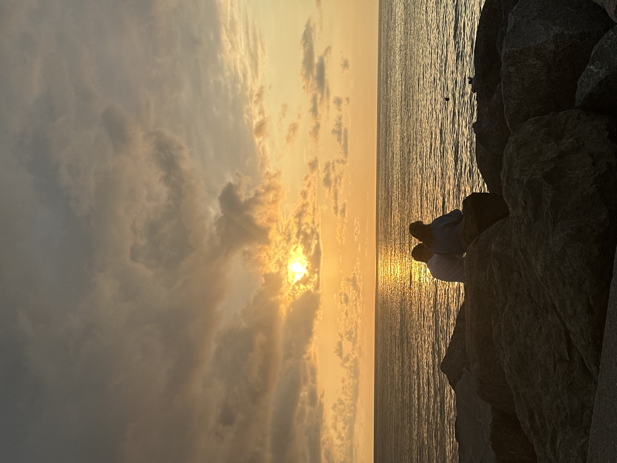 Two people, silhouetted against a dramatic sunset, sit on large rocks near the water's edge. The sky is filled with clouds in shades of orange and gray, with the sun partially obscured by clouds, casting a golden reflection on the rippling sea. The scene is set on a rocky coastline, with the dark, rough texture of the rocks contrasting with the smooth expanse of the water and sky. The location is identifiable as the Batumi Boulevard area in Batumi, Georgia, known for its scenic waterfront and public spaces.