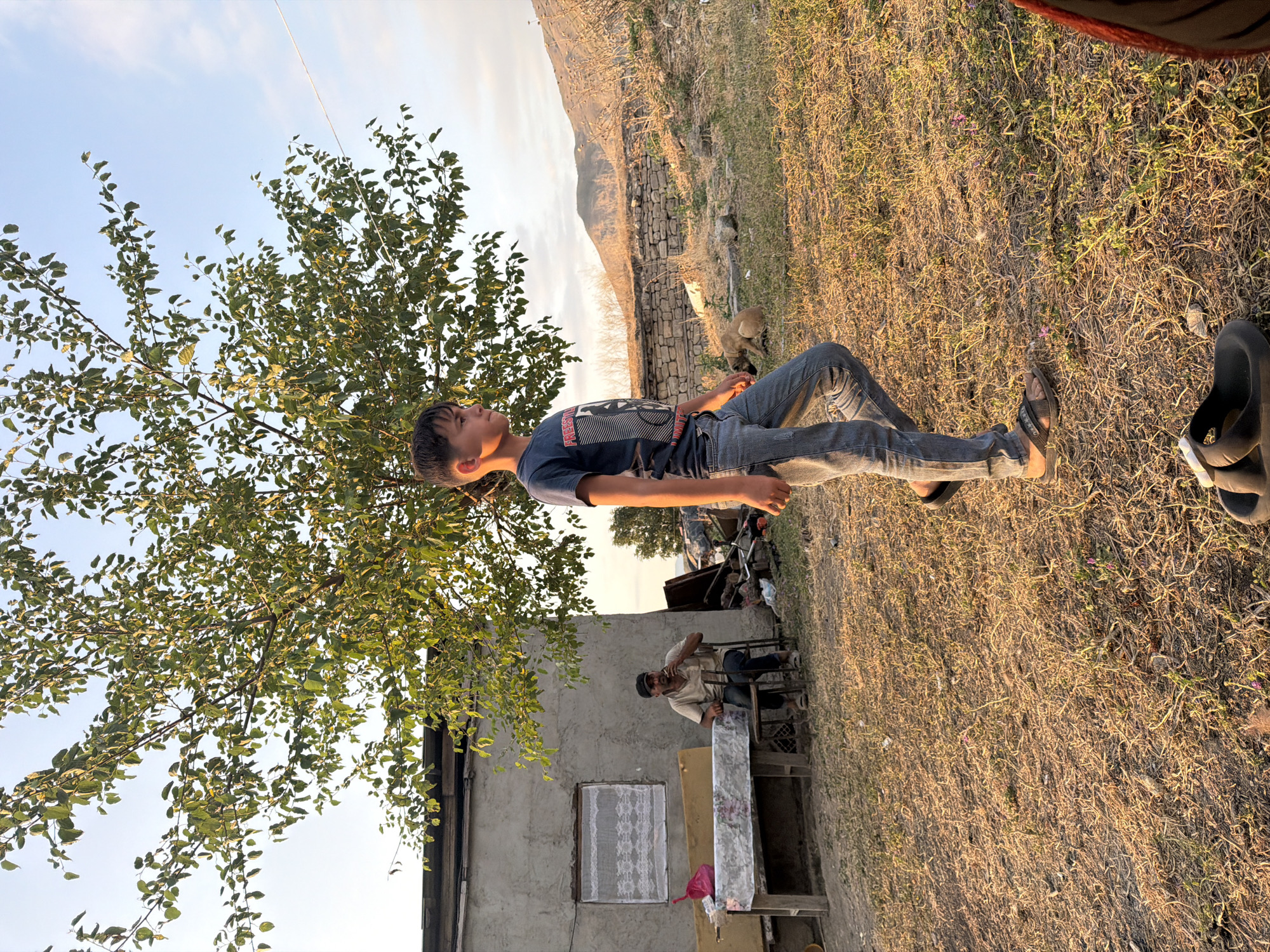 In Köhnə Quşçu, Azerbaijan, a young boy in a dark t-shirt and jeans walks across a dry, grassy field in front of a rustic, concrete building. The building has a single window covered with white lace curtains and a rickety table draped with a patterned cloth is set up outside. An older man, wearing a light-colored shirt and dark pants, sits on a chair at the table, smoking. The scene is set against a backdrop of dry, rocky hills under a bright, clear sky. A small dog is visible lying down in the middle ground.