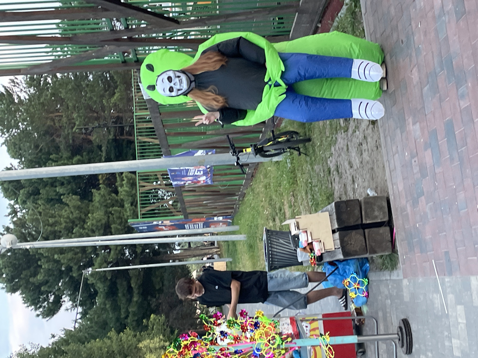 The image depicts an outdoor scene, likely a park or recreational area, in Surgut, Russia. In the foreground, a person is wearing a bright green inflatable alien costume with a skull mask. They are making a peace sign with their left hand. To the left, a young man in a black t-shirt and denim shorts is standing next to a cart filled with colorful pinwheels and other novelty toys. He appears to be attending to the display. Behind the young man is a trash can. Further in the background, a bicycle is parked near a wooden fence. Trees and some advertising banners are visible, suggesting a public space. The ground is paved with brick-like tiles in the foreground and transitions to grass and dirt in the background. The lighting suggests it is late afternoon or early evening.