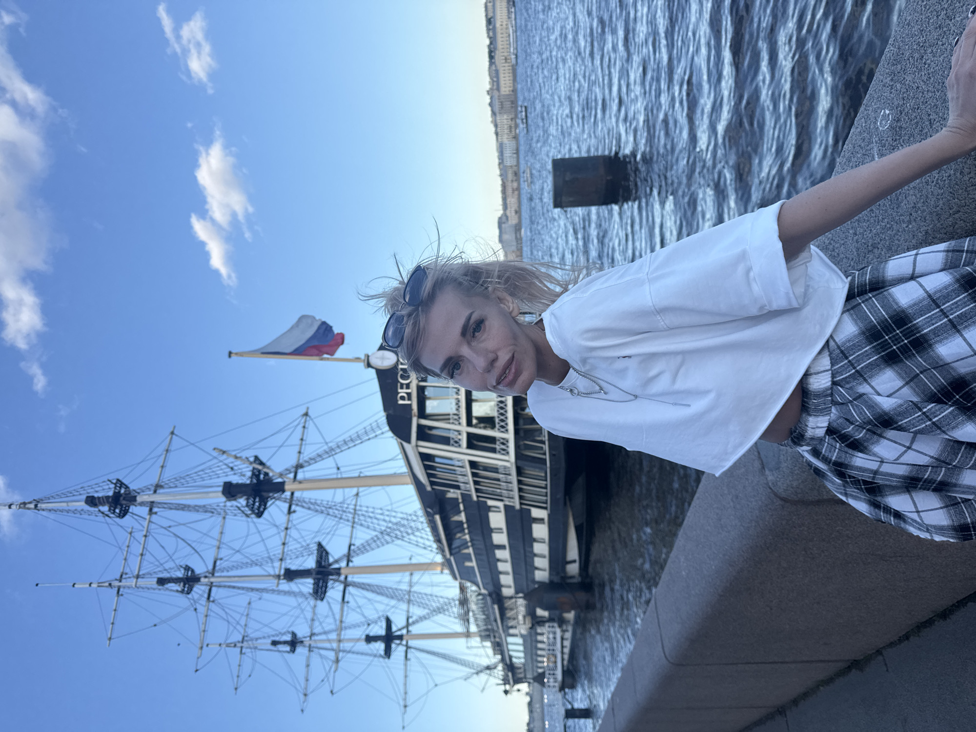 A woman with blonde hair and sunglasses on her head poses for a photo in front of a tall ship docked in a city canal. She is wearing a white t-shirt and a black and white plaid skirt. The ship is black and white with multiple masts and rigging. A Russian flag flies from a pole on the ship. The canal has rippling water, and a stone embankment lines the foreground. In the background, a city skyline with buildings is visible across the water under a clear blue sky with scattered white clouds.