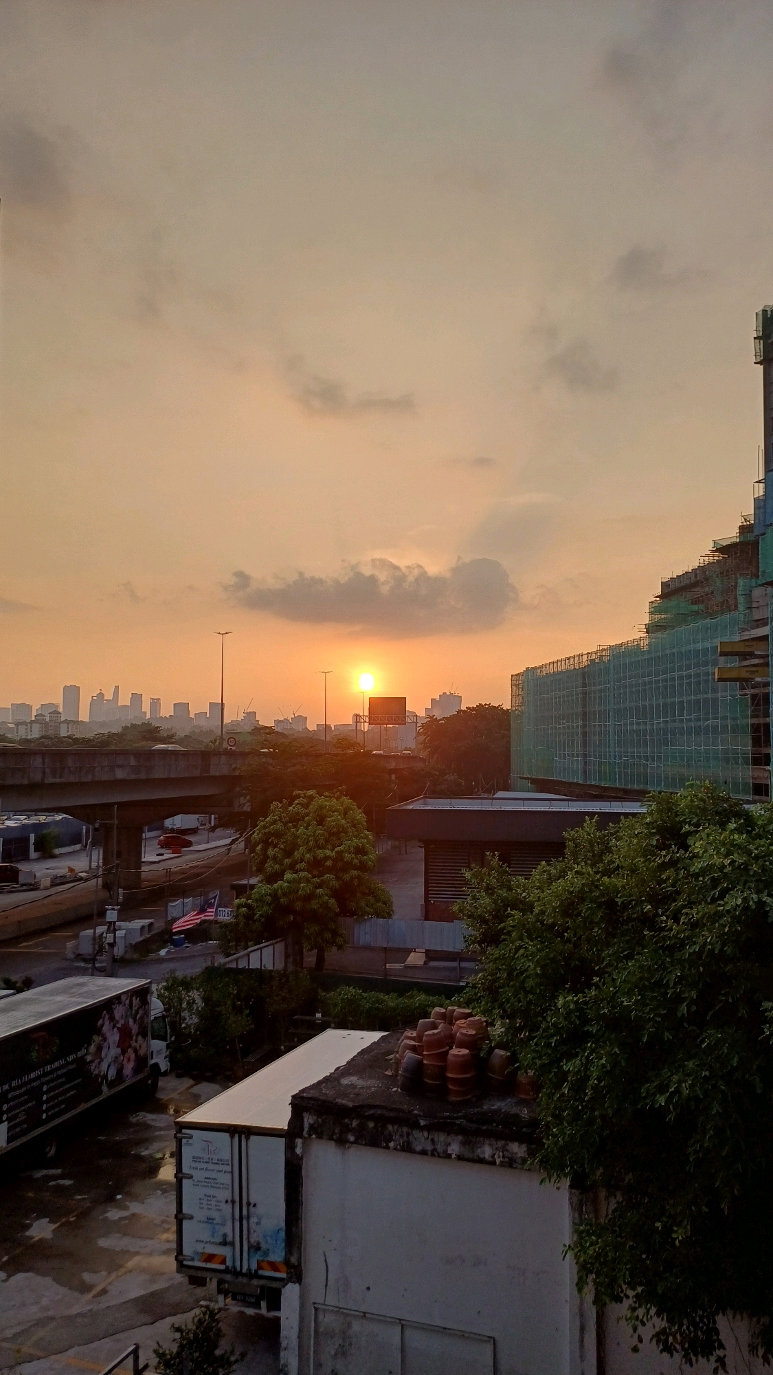 This is a photograph taken during a sunset in Kuala Lumpur, Malaysia. The sun, a bright orb, is visible low in the sky, casting a warm orange glow. In the foreground, there is a construction site with a large building covered in green scaffolding and netting. To the left of the construction site, there is a highway overpass. Several trucks are parked in the lower portion of the image. In the distance, the skyline of Kuala Lumpur is visible, with numerous tall buildings and skyscrapers. The sky is filled with scattered clouds that are illuminated by the setting sun. The overall scene depicts an urban environment during a transitional period of the day.