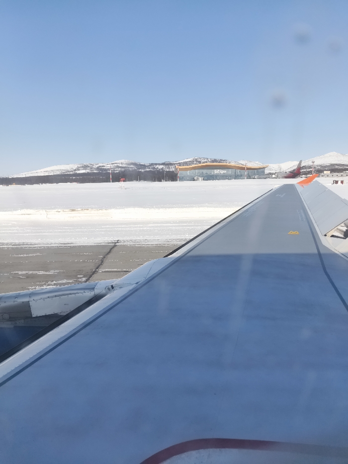 The scene depicts a winter airport environment at Magadan Airport in Sokol, Magadan Oblast, Russia, viewed from an aircraft window. The lower half of the frame is dominated by the light gray right wing of a commercial aircraft, revealing parts of its engine nacelle and an upper surface with yellow markings. The ground below is a snow-covered airfield with visible sections of cleared asphalt runways or taxiways. In the mid-ground, a red conical marker stands on the snow. The background features a modern airport terminal building with a curved, orange-yellow facade and extensive glass windows, prominently displaying the word "МАГАДАН" (MAGADAN) in white Cyrillic lettering. To the right of the terminal, a parked or taxiing aircraft with a red tail and white fuselage is partially visible. Beyond the airport facilities, a dark tree line separates the airfield from distant, snow-covered hills under a clear, bright blue sky. No individuals are visible.