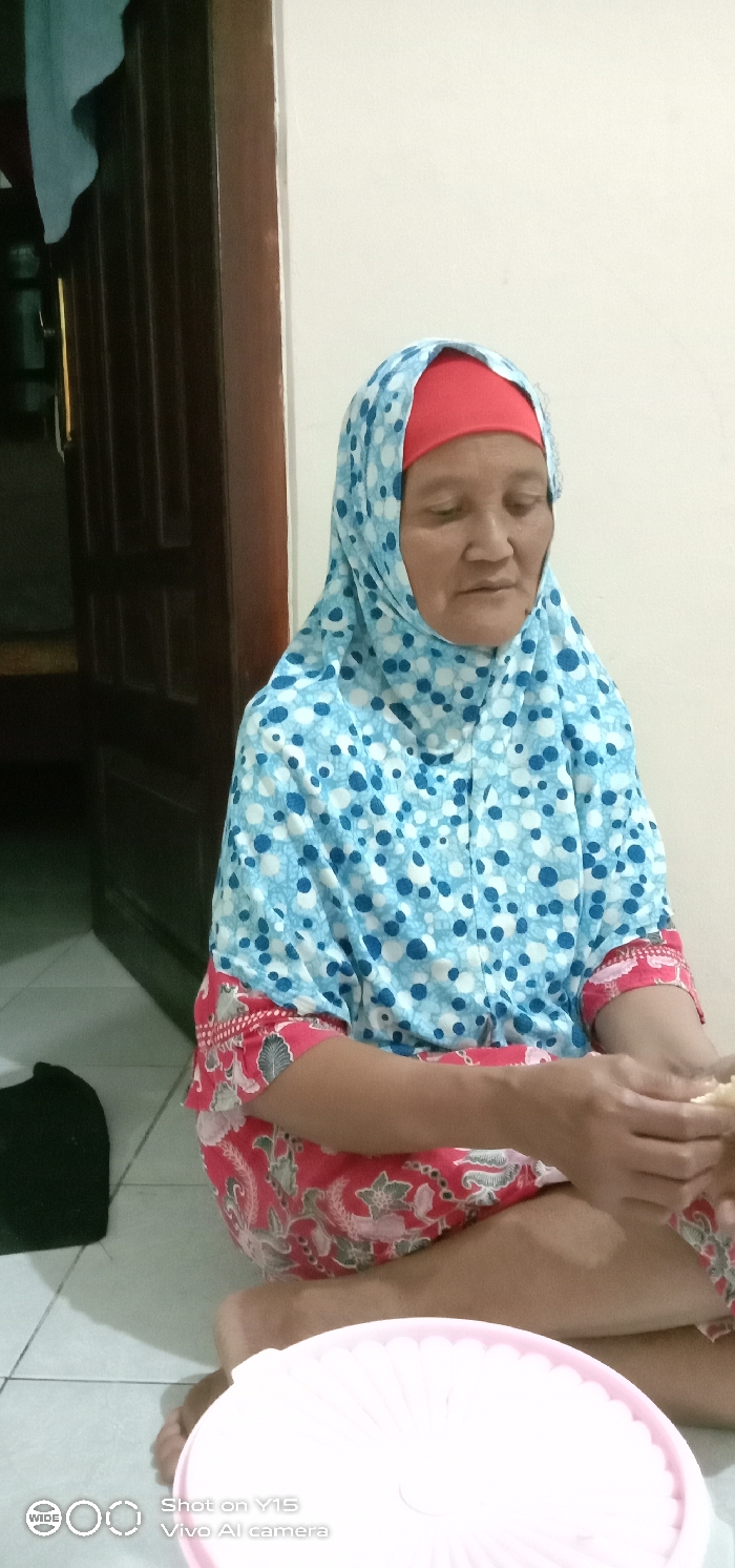An adult female is depicted seated cross-legged on a light-colored tiled floor indoors in Ranugedang, Indonesia. She wears a light blue hijab with white and dark blue polka dots over a red undercap, and a red garment with a patterned design on the sleeves and lower body. Her hands are positioned to hold a small, light-colored, irregular object. Her gaze is directed downwards. A plain light-colored wall forms the background to her right, while a partially open dark brown wooden paneled door is visible to her left. A light pink, round plastic container with a ridged lid is positioned in the lower right foreground. A dark fabric item rests on the floor to the lower left of her bare feet.