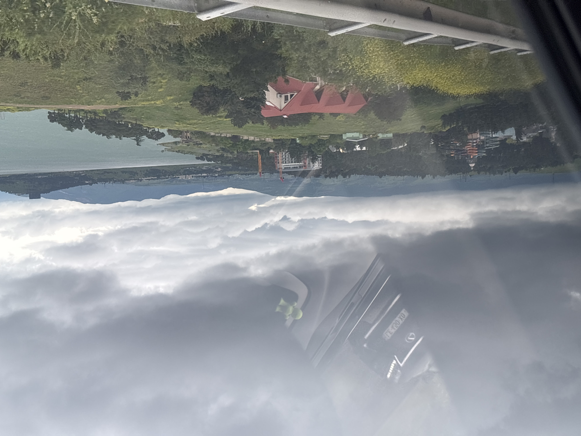 The image depicts a landscape with a body of water (likely a lake or river) in the distance, surrounded by greenery and buildings. There's a house with a red roof visible in the middle ground. In the foreground, a section of a vehicle's interior is visible, with a dashboard and possibly a windshield obscured by clouds or fog. The license plate of the car is visible, reading "TL 020 TL". The overall scene appears to be taken from a high vantage point, possibly overlooking the landscape. The location is possibly in the city of Tbilisi, Georgia.