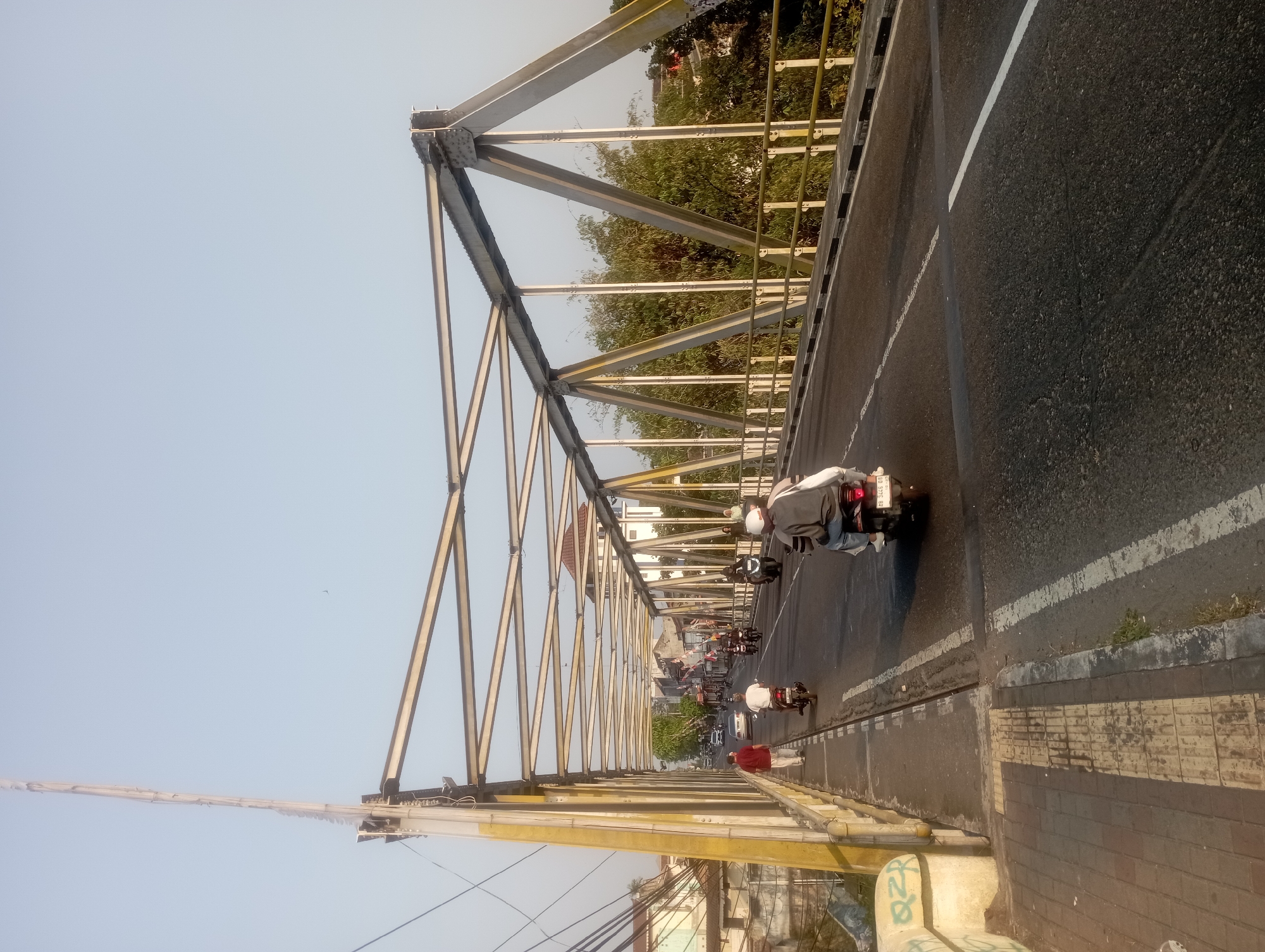 The image shows a view of a bridge from the perspective of a person standing on the sidewalk. The bridge has a steel truss structure and is painted yellow and grey. The roadway on the bridge is asphalt and has white lane markings. There are several people on motorcycles, a car, and two pedestrians visible on the bridge. The weather appears to be clear and sunny. The scene suggests an urban environment, likely in Yogyakarta, Indonesia, given the architecture and the presence of motorcycles as a common mode of transportation. The bridge itself seems to be an older structure, possibly a historical landmark. The surrounding area shows buildings and trees.