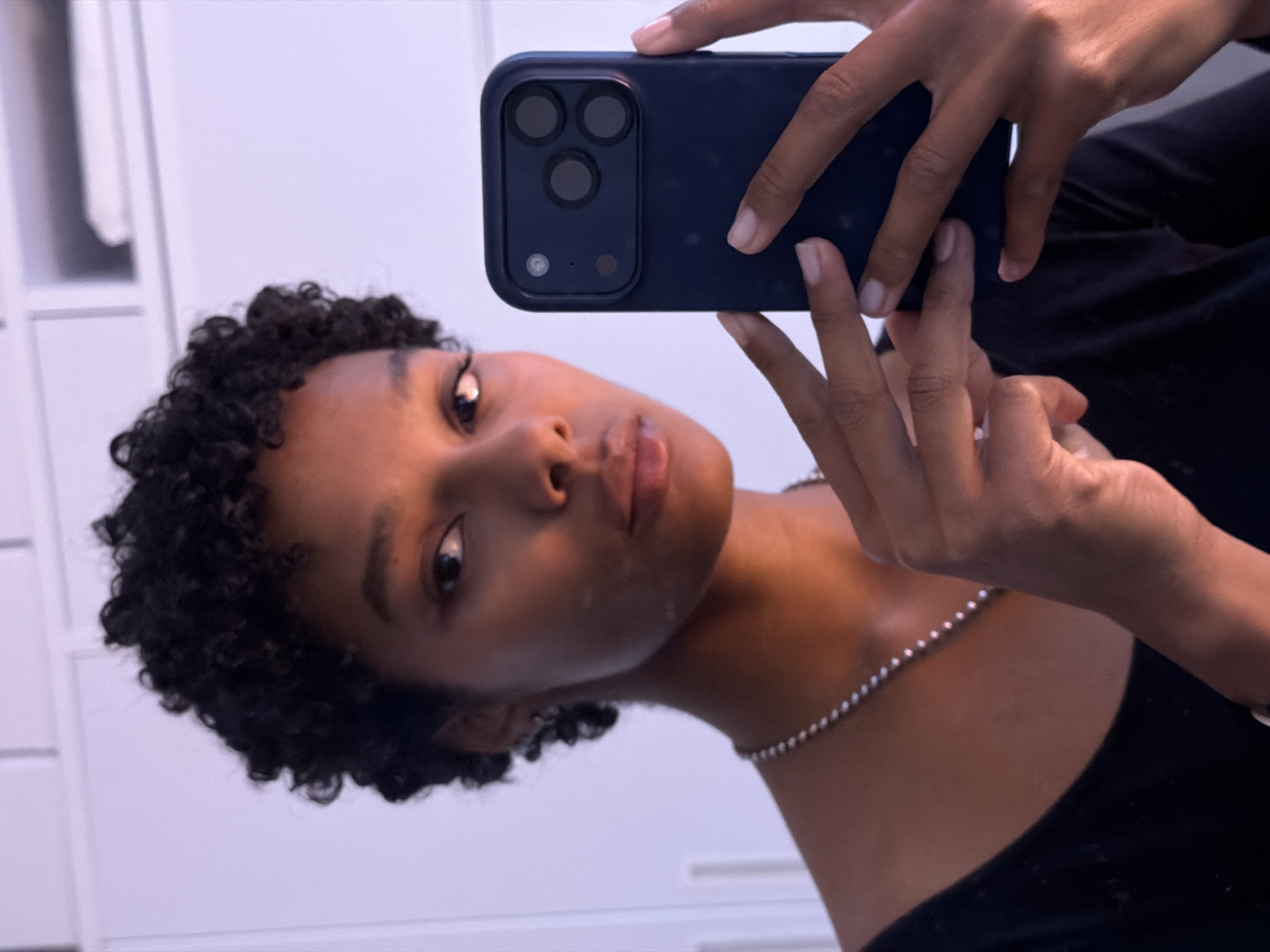 A young Black woman with short curly hair takes a selfie in a bathroom. She holds a dark-colored smartphone in front of her face, her fingers visible on the back and sides of the device. She wears a dark top and a delicate silver necklace. The background includes a white door or cabinet with horizontal paneling. The location cannot be determined as Paris, France, as no evidence of the city is present.
