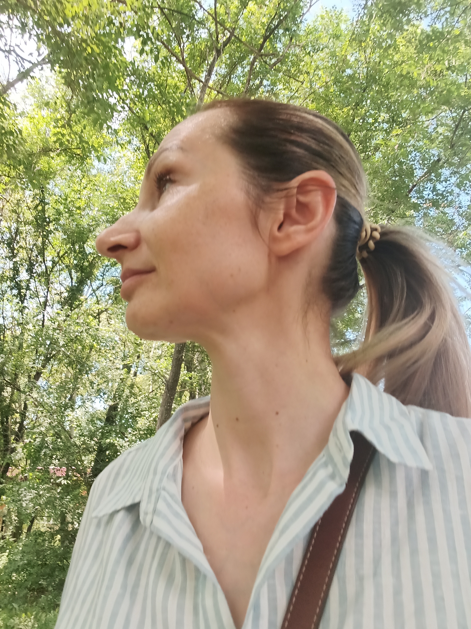 A medium shot captures a woman in profile, looking to her left against a backdrop of lush green trees and a clear blue sky. She has fair skin, brown hair pulled back in a ponytail with a light brown elastic, and is wearing a light blue and white striped collared shirt. A brown strap is visible across her shoulder, suggesting she is wearing a bag. The lighting indicates it is daytime, likely sunny, with dappled sunlight filtering through the leaves. The scene appears to be in an outdoor, natural setting, such as a park or wooded area. Based on the provided information, the location is Komsomolsk-on-Amur, Russia.