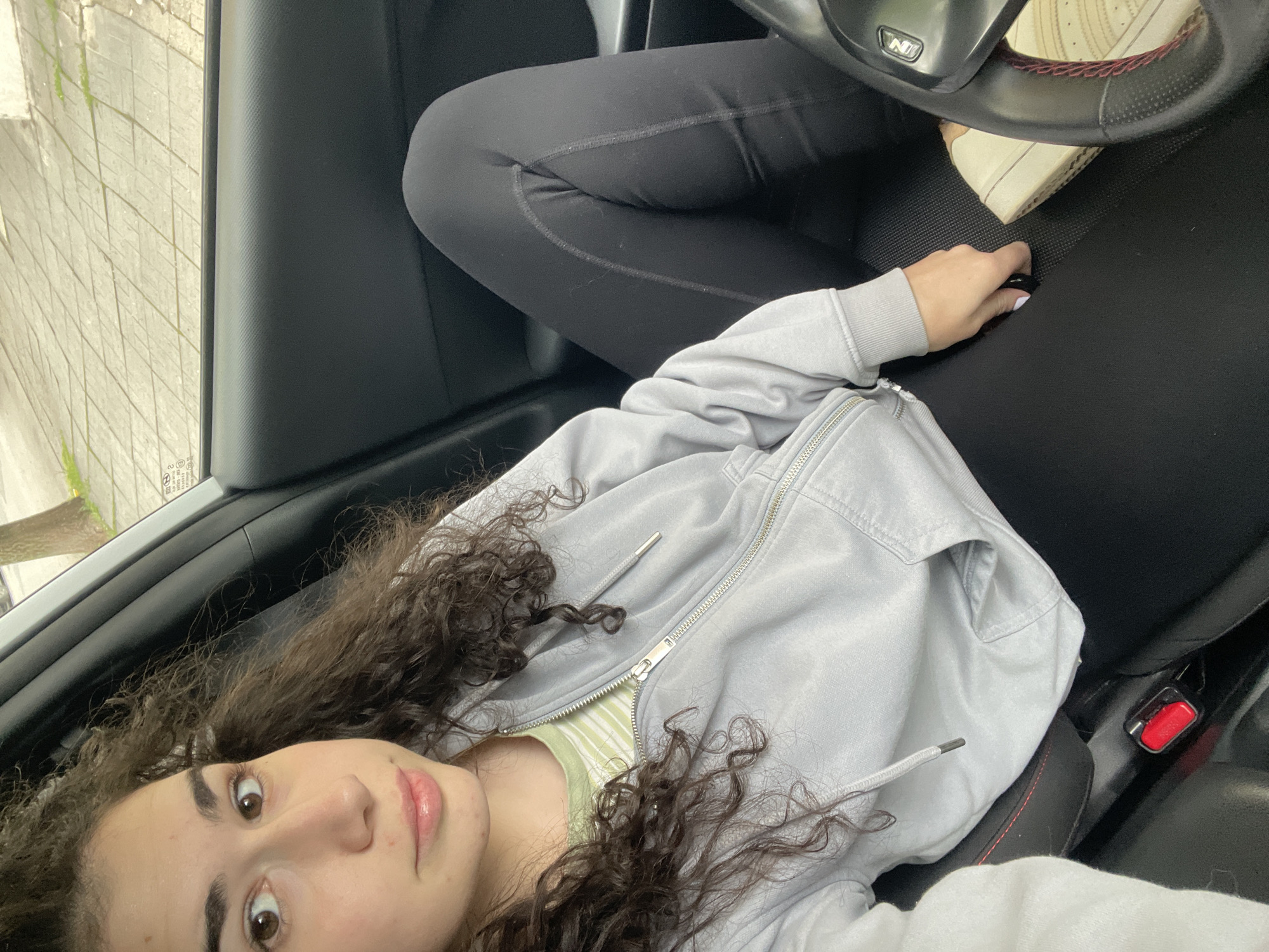 A young woman with curly brown hair is sitting in the driver's seat of a car. She is wearing black leggings and a light gray zippered hoodie over a striped shirt. Her legs are up on the dashboard, and her right foot, wearing a beige sneaker, is near the steering wheel. Her right hand is near the steering wheel. The car is parked near a sidewalk. Outside of the car window, there are glimpses of buildings and greenery, possibly trees. The specific location in Sofia, Bulgaria is not discernible from the image.