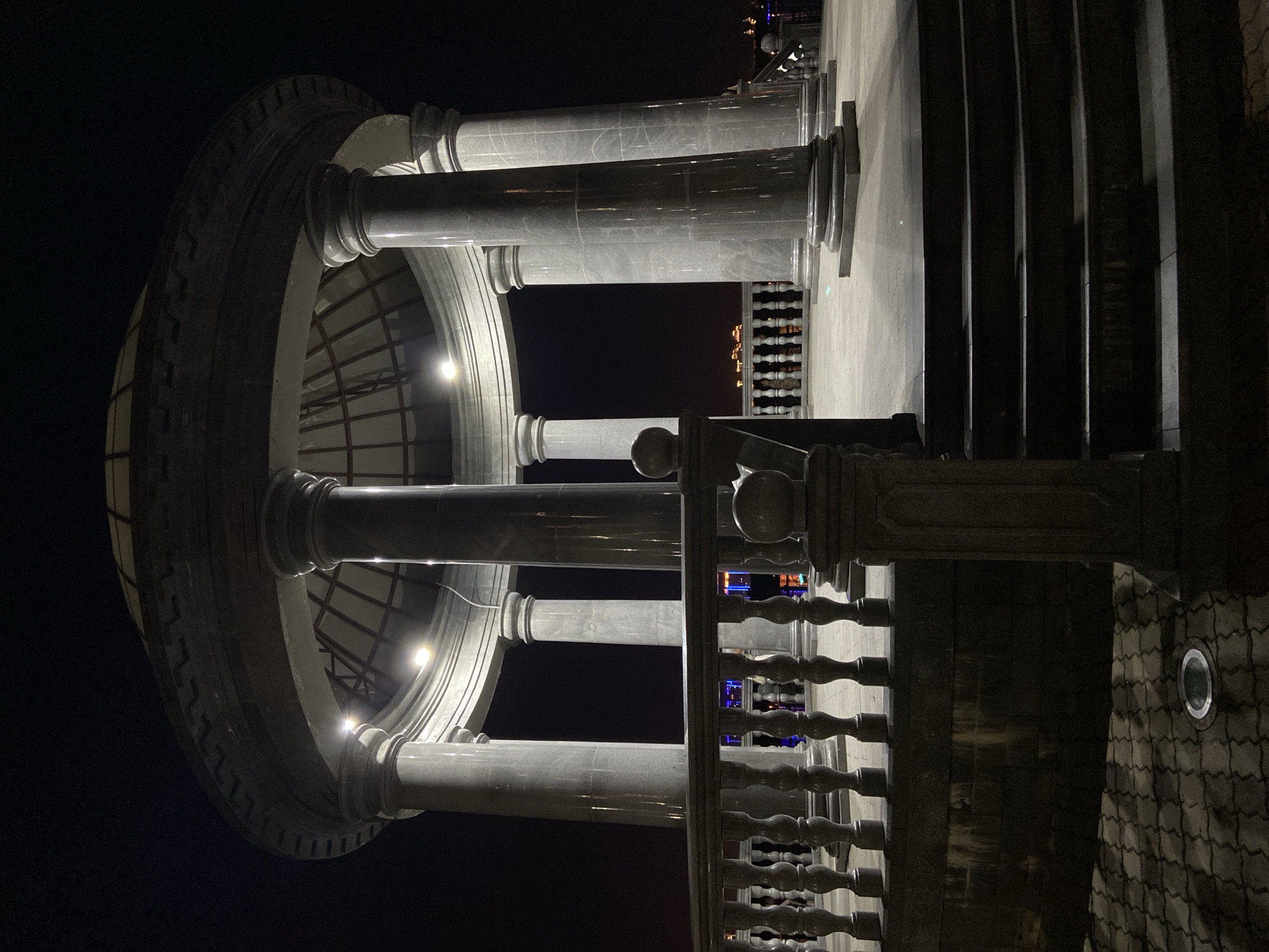 The image depicts a rotunda at night, likely a public structure, possibly in Blagoveshchensk, Russia, given the provided context. The rotunda features a domed roof supported by several large, marble columns. A decorative railing runs along the edge of the platform. In the background, the lights of a city are visible, suggesting an urban setting. The ground in the foreground is paved with interlocking bricks, and a set of stairs leads up to the rotunda. There are no people visible in the image. The lighting is artificial, with lamps illuminating the rotunda and the distant city lights providing a backdrop. The overall scene is dark, with shadows cast by the architecture.
