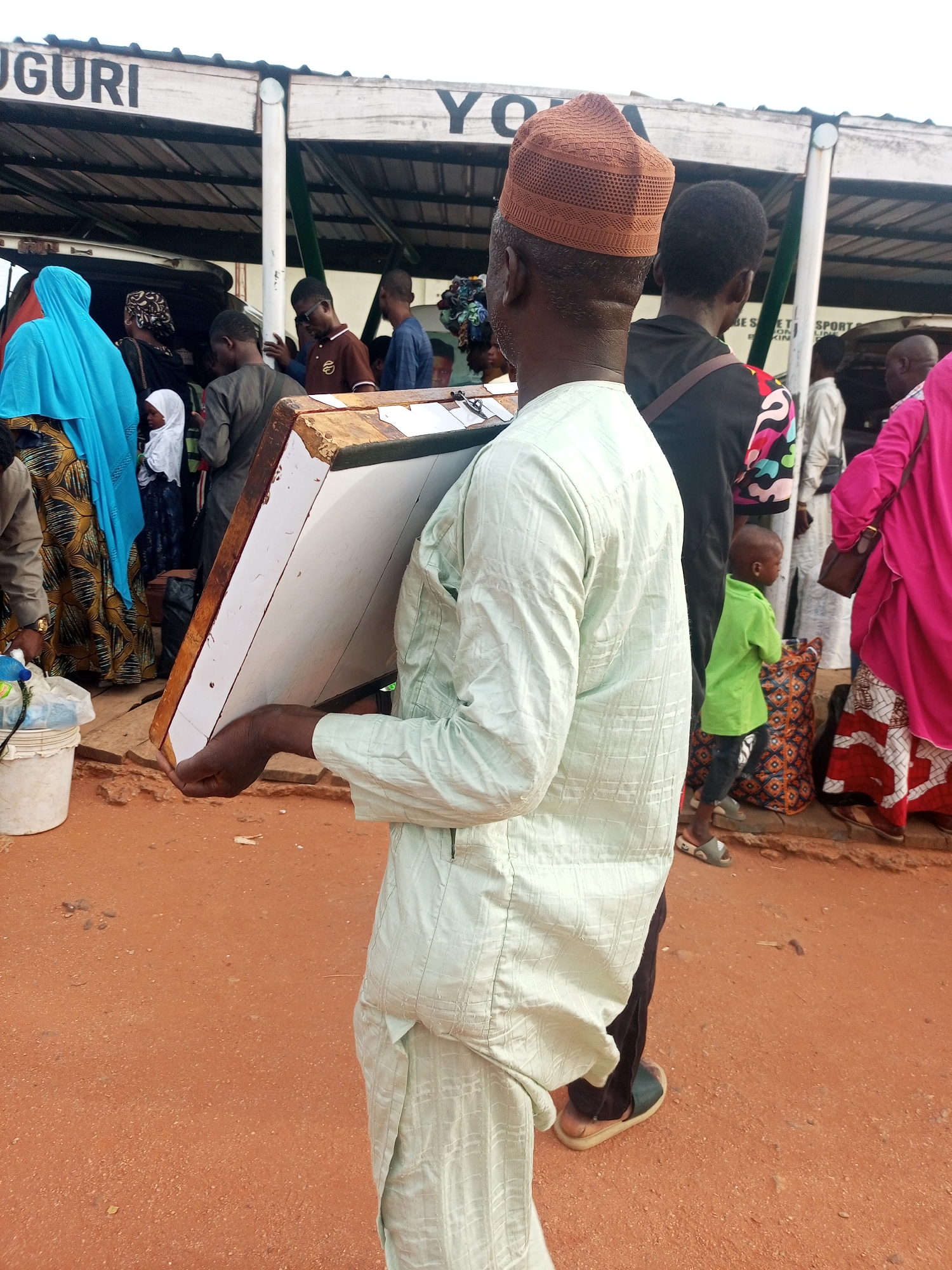 In Gombe, Nigeria, a man wearing a light green robe and a brown kufi cap carries a wooden box with a white interior surface. He is among a crowd of people, some wearing traditional garments. A sign above reads "GURI YO". The ground is reddish-brown.