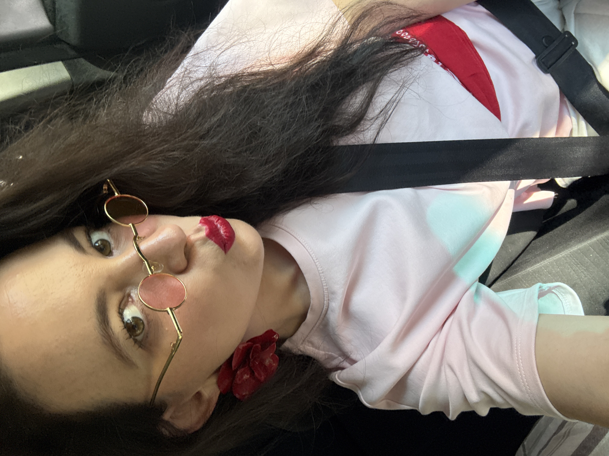 A close-up, selfie-style shot features a woman in a light pink t-shirt with a red design on the right side. She is wearing round, gold-rimmed sunglasses with pink lenses and large, red, flower-shaped earrings. Her lips are painted a bright red. A black seatbelt is visible across her chest. The background is dark, suggesting the interior of a vehicle. There is no discernible information about the location in Chaikovsky, Russia.