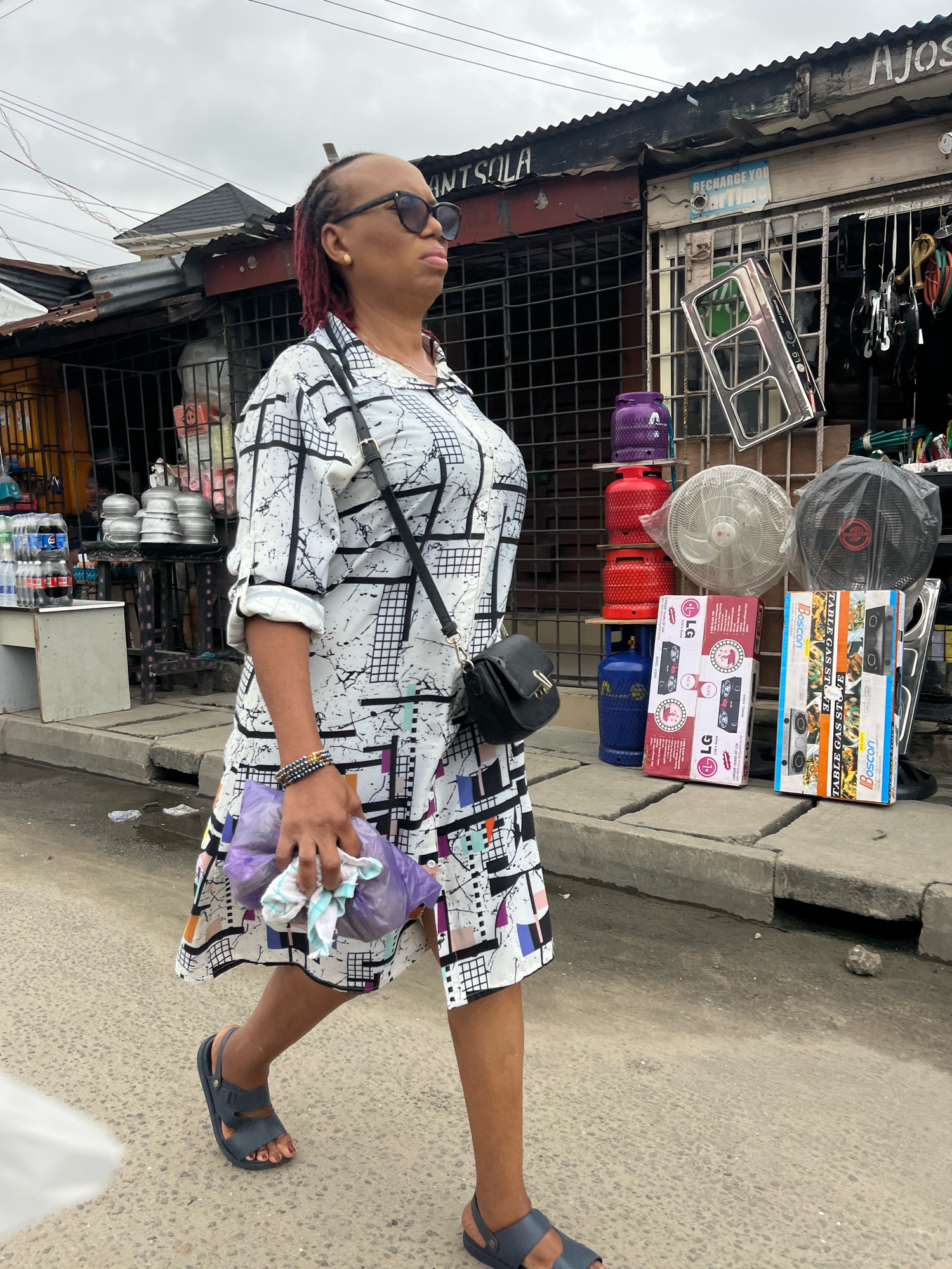 In Ado, Nigeria, a woman with red-dyed braided hair walks from left to right in front of a row of market stalls. She wears sunglasses, a black crossbody bag, and a white dress with a black geometric pattern. In her right hand, she carries a purple plastic bag with white cloth items. The stalls display various goods, including stacked gas cylinders (one blue, one red, one purple), electric fans, and boxes of kitchen appliances. A metal grate covers a portion of one stall, and a sign reading "AJOS" is visible above. The ground is paved with asphalt, and a concrete drainage ditch runs along the curb. The sky is overcast.