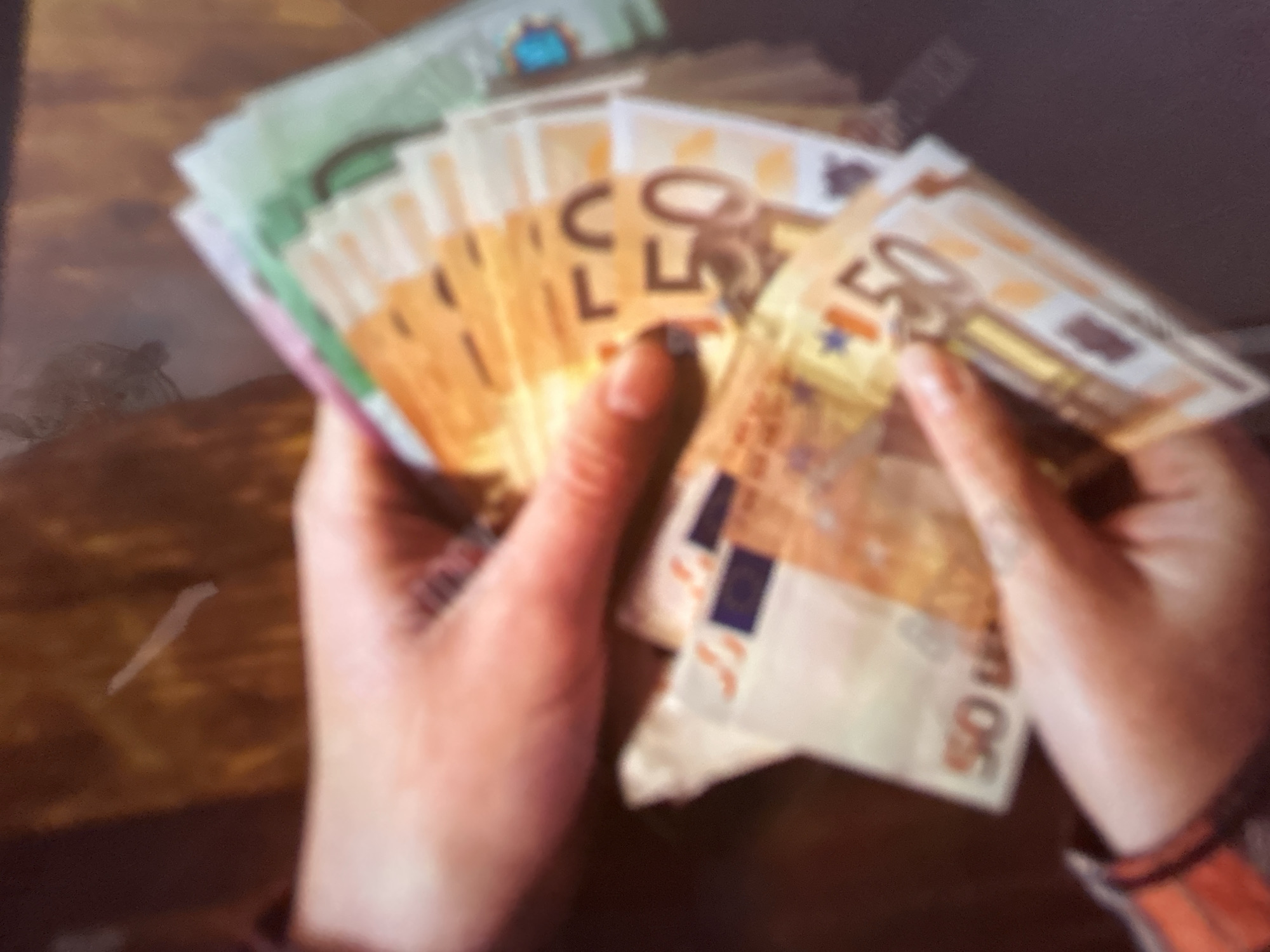 The media file shows a close-up, blurry image of a person's hands holding a fan of Euro banknotes. The banknotes are predominantly yellow and orange, with some green and white visible at the edges, suggesting denominations like 50 Euro notes. The hands are holding the money fanned out, with some fingers extended and others curled, indicating the act of counting or displaying the cash.
The background is dark and out of focus, with a wooden surface visible beneath the hands. A faint, elongated white mark is also present on the wooden surface. The lighting is warm, casting a slight glow on the money and hands. There are no clear indicators of time of day, weather, or specific emotions, beyond the implied focus on handling money.
There is some visible text on the banknotes. The number "50" is clearly legible on several notes. Other text is blurry but appears to be in a European language, consistent with Euro currency. The overall impression is one of wealth or financial transaction.