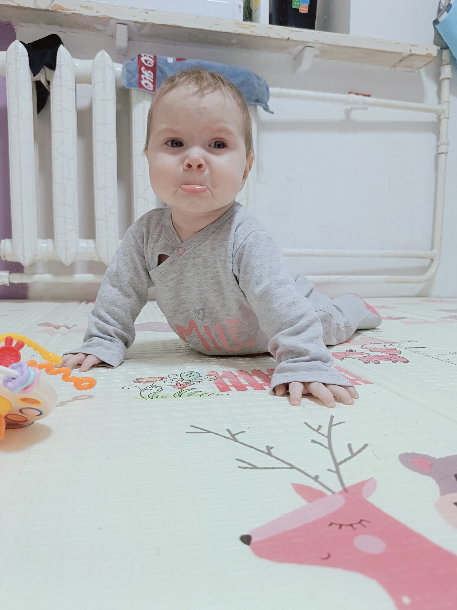 A baby wearing a grey pajama set is lying on its stomach on a light-colored playmat decorated with cartoon animals, including a pink deer. The baby is in the foreground, with its head raised and tongue slightly protruding. To the left is a toy, and in the background is a white radiator. The location is most likely inside a residence in Rusalevka, Russia, given the provided information.