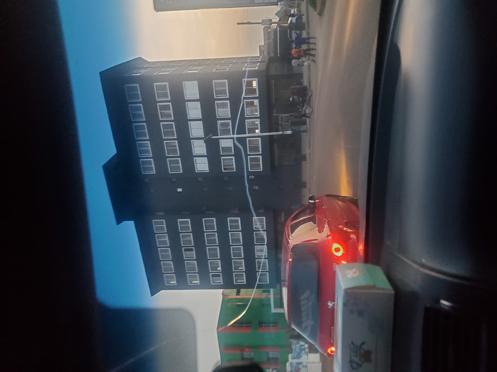 The image captures a view from inside a vehicle in Sardauna, Nigeria, showing a street scene at dusk. A dark, modern, multi-story building with numerous rectangular windows dominates the background. To the left, a green building with orange vertical accents is visible. In the foreground, the rear of a red Peugeot hatchback is prominent, with its taillights illuminated. A tissue box rests on the dashboard in the lower left corner. Several figures are seen walking and standing on the street near the dark building, and a motorcycle is parked in the vicinity. The sky is a soft, hazy gradient of orange and blue, indicating either sunset or sunrise.