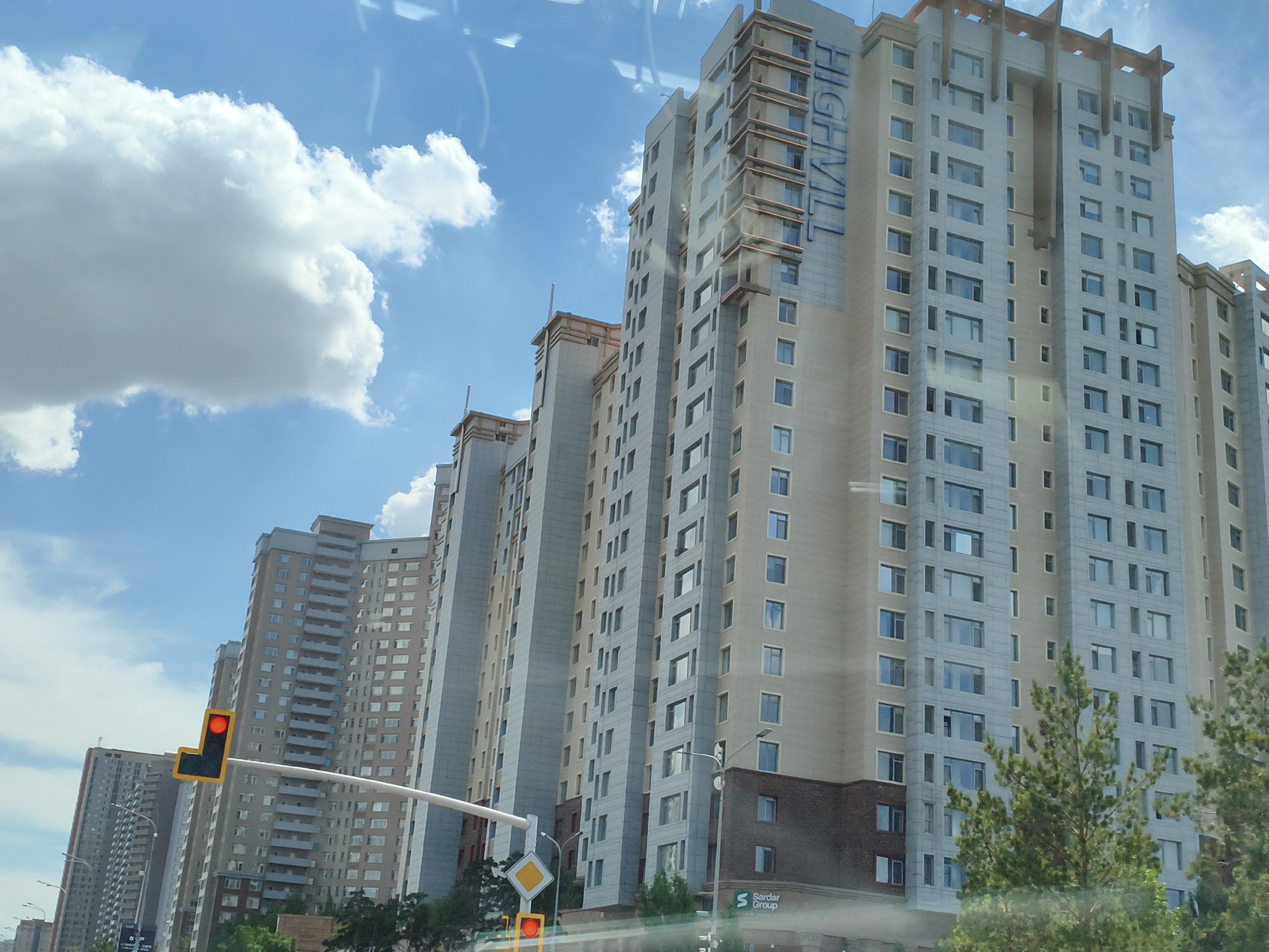 This is a medium-range, eye-level, outdoor shot taken on a bright, partly cloudy day in Astana, Kazakhstan. The image features a row of tall, modern, cream-colored residential apartment buildings with many windows and balconies. The buildings are arranged in a receding line, creating a sense of depth. On the left side of the frame, a traffic light with a red signal is visible, suspended by a white curved pole. In the foreground, lush green trees partially obscure the lower portions of the buildings. The sky is a clear blue with scattered white clouds. The overall scene depicts a typical urban environment in Astana, with a focus on the residential architecture and infrastructure.