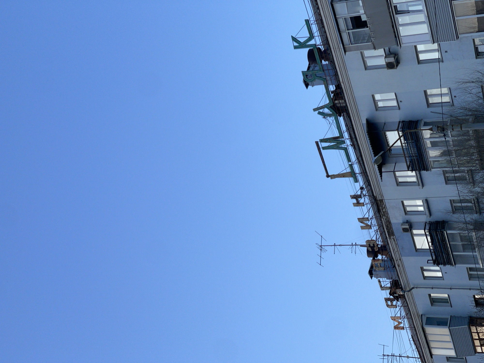 The image captures a clear blue sky occupying the majority of the left side of the frame, contrasting with a multi-story residential building dominating the right side. The photo appears to be rotated 90 degrees clockwise, giving a perspective as if looking upwards at the building's facade.
The building is an apartment block with a light grey or pale blue exterior, featuring numerous rectangular windows, some of which have enclosed balconies. Several external air conditioning units are mounted below windows. Bare tree branches are visible along the far right edge, suggesting either winter or early spring in Krasnoyarsk, Russia.
Prominently displayed along the upper portion of the building's facade is large, blocky Cyrillic text, rendered in a teal or green color, mounted on a metal frame. The clearly discernible letters spell out "ЖКХ МИР" (ZhKKh MIR), which translates to "Housing and Utilities Services 'Peace'" or "'World'". There are other fragmented letters further along the sign that are difficult to fully decipher. Multiple television or radio antennas are visible on and around the building's upper structure.
The scene depicts a static urban landscape under bright, clear daylight, indicating good weather. There is no specific activity or event taking place. The architectural style and Cyrillic script are consistent with the provided location context of Krasnoyarsk, Russia.