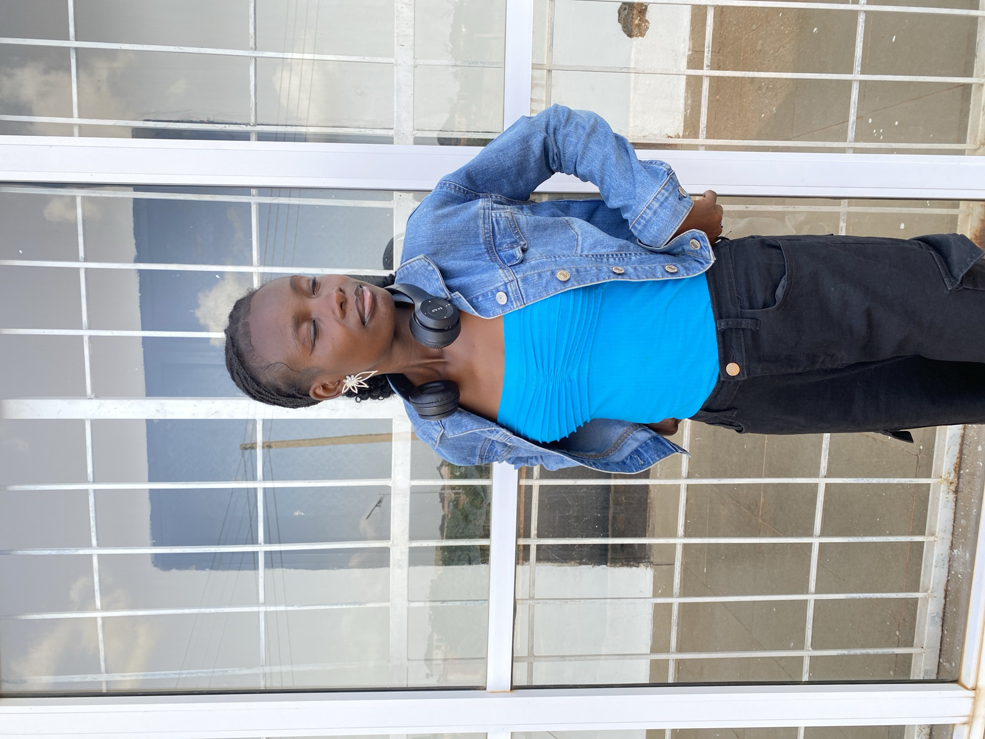 A young Black woman with braided hair stands in front of a windowed building in Bar Jirgi Summa, Nigeria. She is wearing a blue, strapless, ruched top, a denim jacket, and black cargo pants. Black headphones are around her neck, and she has large, star-shaped earrings. Her eyes are closed, and her head is tilted slightly. The background shows a reflection of the sky and some buildings in the window panes, which are separated by white vertical and horizontal bars. The lower part of the building appears to be made of stone.