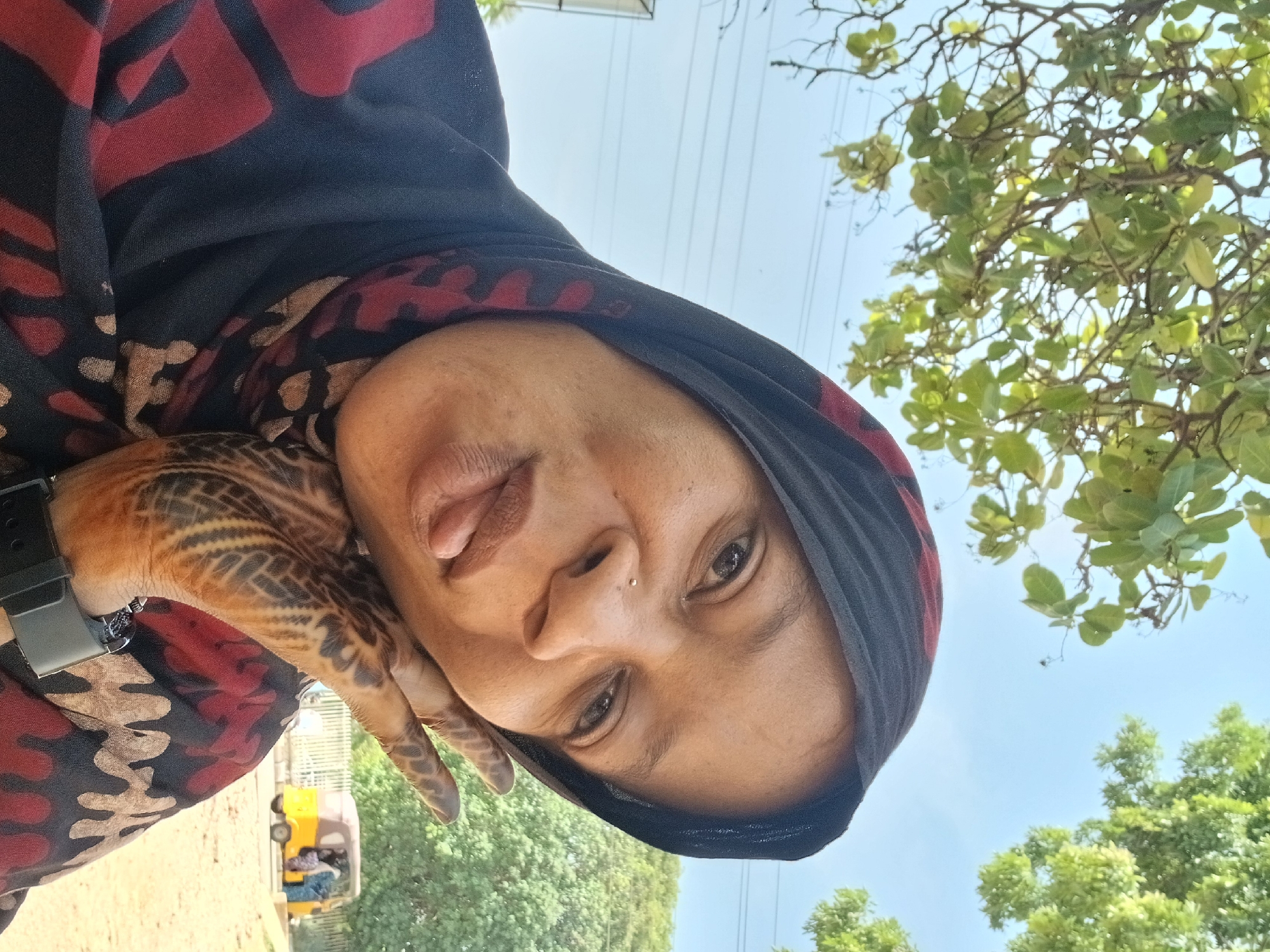 This outdoor portrait features an individual, likely a woman, looking directly at the camera with her head tilted to the left. She is wearing a black head covering, possibly a hijab, patterned with bold red and earthy-toned abstract shapes. Her complexion is warm, and she has a small nose stud on her right nostril. Her expression appears calm, with her tongue slightly visible.
Her right hand, positioned near her chin, displays intricate, dark brown mehndi or henna designs across the back of her hand and fingers, featuring geometric and possibly floral patterns. A dark wristwatch is visible on her left wrist.
The background is bathed in bright daylight under a clear blue sky, indicating fair weather. Lush green trees and foliage are visible in the upper right and lower left parts of the frame. Faint utility lines can be seen strung across the sky. In the blurred lower left background, a yellow auto-rickshaw (often referred to as a tuk-tuk) is partially visible, suggesting an urban or semi-urban street setting. The image itself is rotated, giving the composition an angled perspective. The presence of the auto-rickshaw and the overall environment align with a location like Shaba, Nigeria. No discernible text is visible in the image.