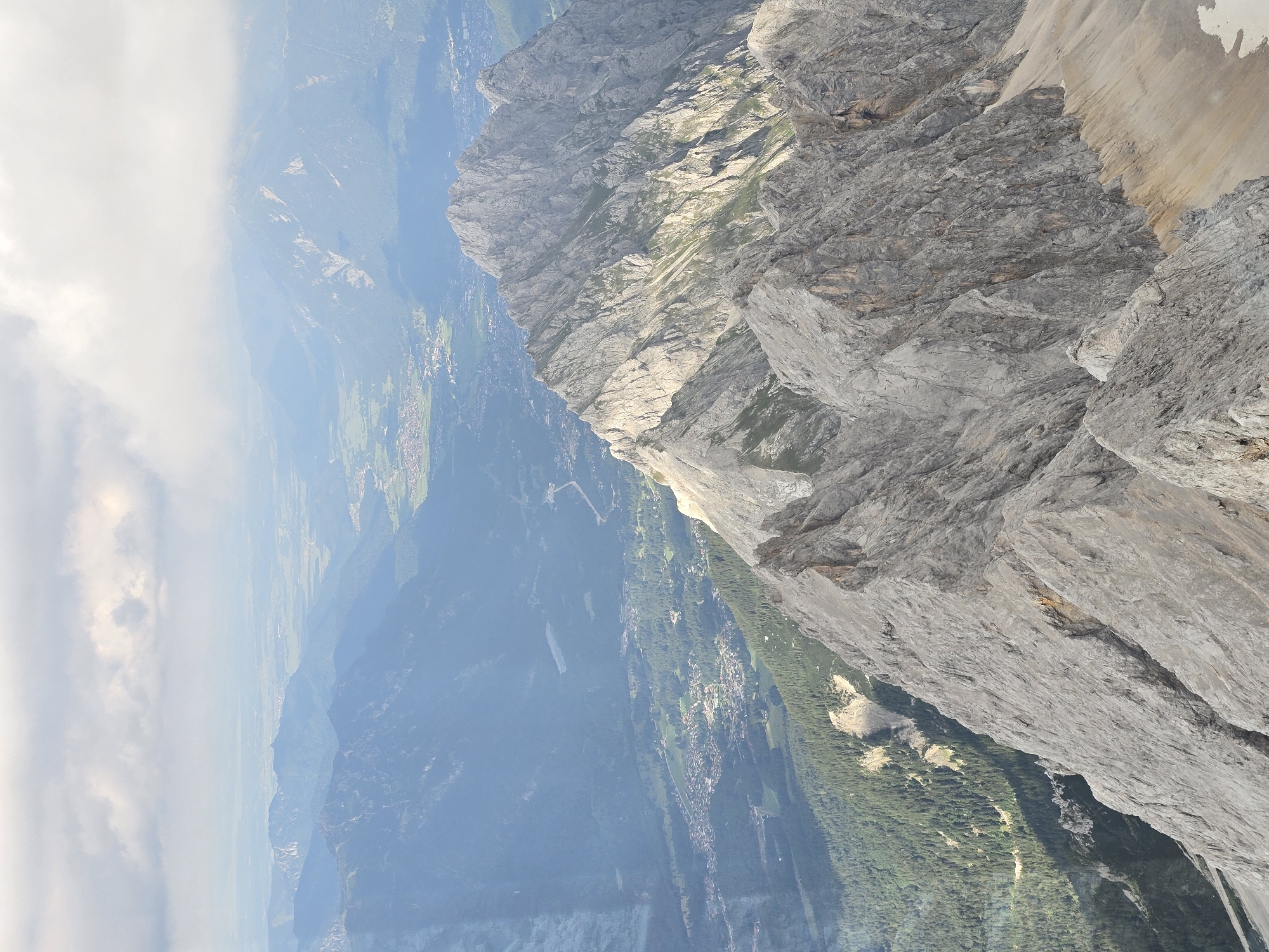 This media file captures a dramatic, vertically oriented view of a mountainous landscape. The foreground is dominated by the rugged, gray rock face of a mountain, with sharp inclines and textured surfaces that suggest steep drops and geological formations. Sunlight casts shadows across the rock, highlighting its contours and creating a sense of depth.
Beyond the immediate mountain, the scene unfolds into a vast panorama of valleys and lower mountain ranges. Patches of green forests and fields are visible, interspersed with what appear to be small settlements or villages, characterized by clusters of buildings. The distant landscape is softened by a hazy, bluish atmosphere, common in mountain environments, and hints at further mountain peaks fading into the background.
Clouds are present, both in the distance, forming a band of white against the blue sky, and closer to the foreground, partially obscuring some of the mountain peaks. The lighting suggests it is daytime, likely mid-morning or afternoon, given the angle of the sun.
The overall impression is one of grand scale and natural beauty, emphasizing the imposing presence of the mountains and the expansive vista they overlook. There is no discernible activity or people in the image, focusing solely on the natural environment. The location context of Grainau, Germany, suggests this could be a view from the Zugspitze, Germany's highest peak, known for its breathtaking alpine scenery. No text is visible in the image.
