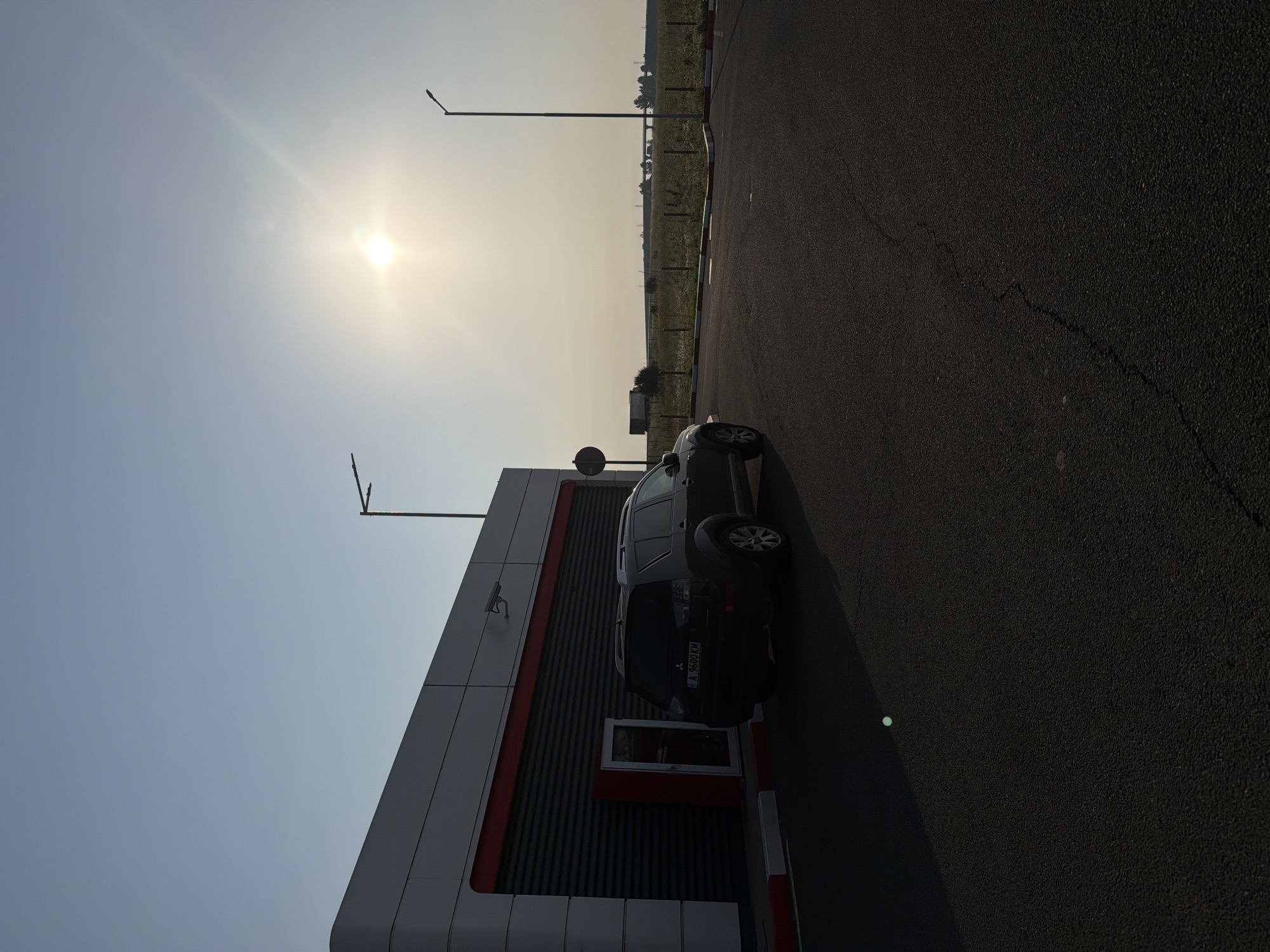 The image, when viewed in correct landscape orientation, captures a bright, sunny daytime scene featuring a modern commercial building, a dark-colored SUV, and a vast open field adjacent to a paved area.
In the upper left portion of the image, the sun shines brightly against a clear, light blue sky, casting a strong glare and long shadows, indicating either morning or late afternoon. Several tall streetlights are silhouetted against the sky, with one prominent pole extending upwards from the middle of the frame.
To the left, a contemporary building with gray paneling and a horizontal red stripe stands prominently. The building features vertically ridged sections and a window with a red frame. A security camera is mounted on the building's exterior. Parked in front of the building, with its rear facing the viewer, is a dark SUV or crossover, distinguished by a spare tire mounted on its tailgate. The car's license plate is clearly visible and reads "A 9600 KM", indicating it is a Bulgarian vehicle (Sofia region code 'A').
The foreground and midground on the right side of the image are dominated by an asphalt surface, likely a parking lot or a section of a road. Red and white curb markings delineate the edge of the paved area near the building. A distinct crack runs diagonally across the asphalt in the lower right. Beyond the paved area, a long stretch of green and golden field, possibly farmland, extends into the distance, bordered by a fence. Faint structures, possibly a distant bridge or other buildings, are visible on the horizon under the bright sky.
The scene depicts a static moment at what appears to be a roadside stop or service area in a semi-rural setting near Vodenichane, Bulgaria. There are no people visible, and the overall impression is one of calm and stillness under clear, sunny weather.