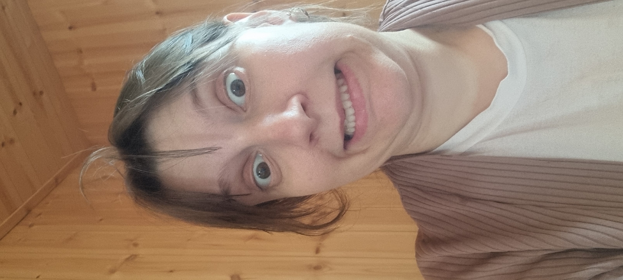 A fair-skinned woman with light brown hair and wide eyes smiles broadly in a close-up, slightly distorted self-portrait. Her head is tilted to the left, with the wood-paneled ceiling and floor visible in the background. She is wearing a white t-shirt and a light brown, horizontally ribbed cardigan. The location appears to be indoors, likely a private residence given the wooden paneling. There is no evidence indicating this image was taken in Dunnae-myeon, South Korea.