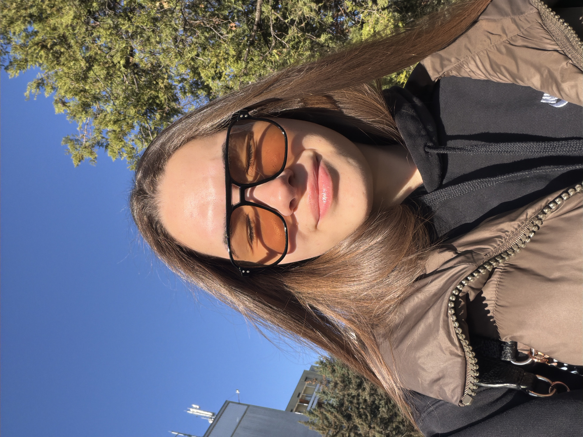 A selfie of a young woman with long, straight brown hair wearing black framed, amber-tinted sunglasses. She is wearing a brown puffer jacket over a black hooded sweatshirt. The background shows a blue sky, green tree foliage, and a building with antennas visible in the lower left corner. The location is likely Sofia, Bulgaria.
