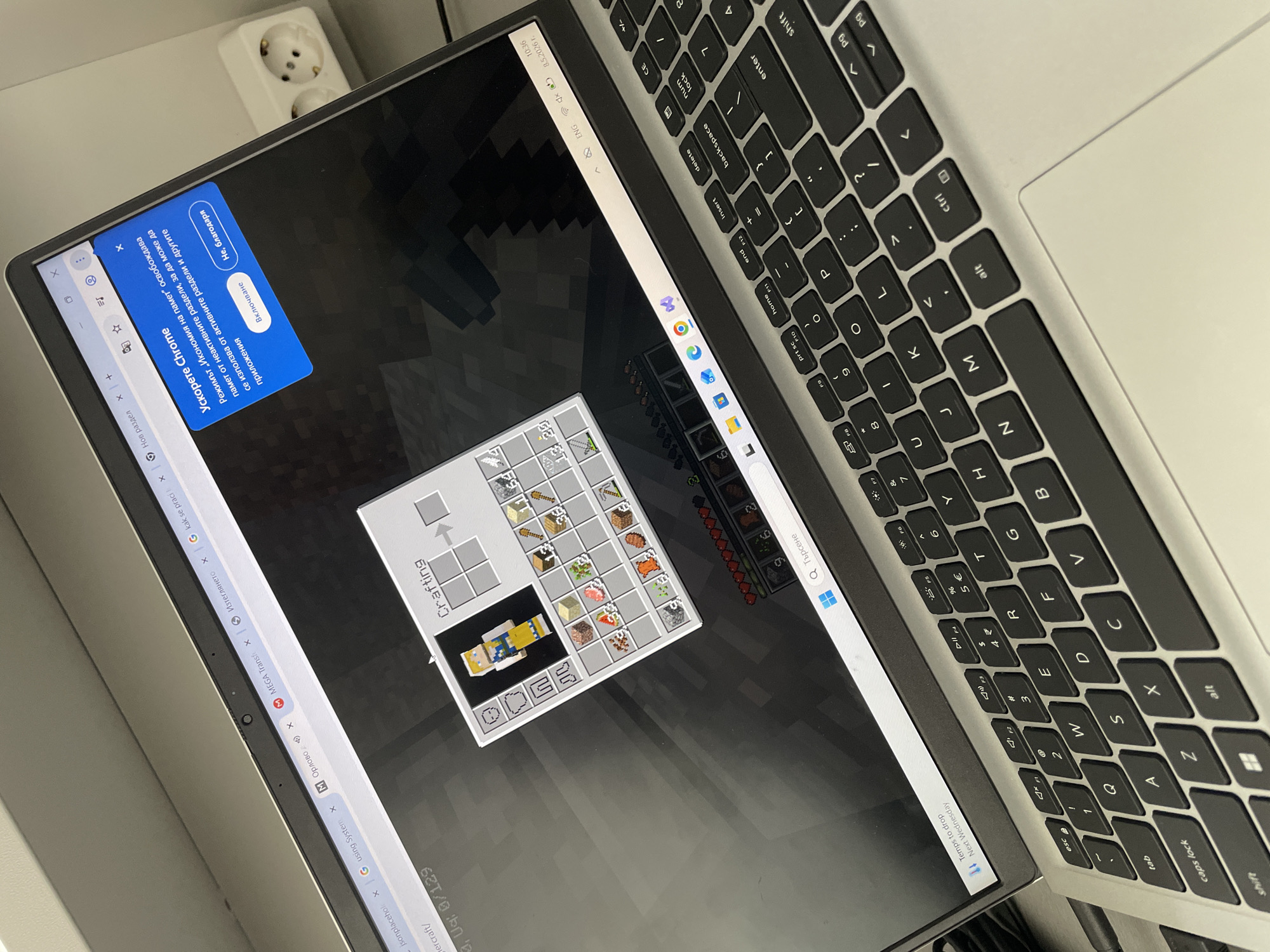 A laptop is open, displaying a Minecraft game. The crafting menu is visible with materials placed for a character skin. A Chrome browser with multiple tabs is open in the background, with some tabs labeled in a Cyrillic language. A keyboard sits to the right of the screen. A two-plug electrical outlet is nearby.