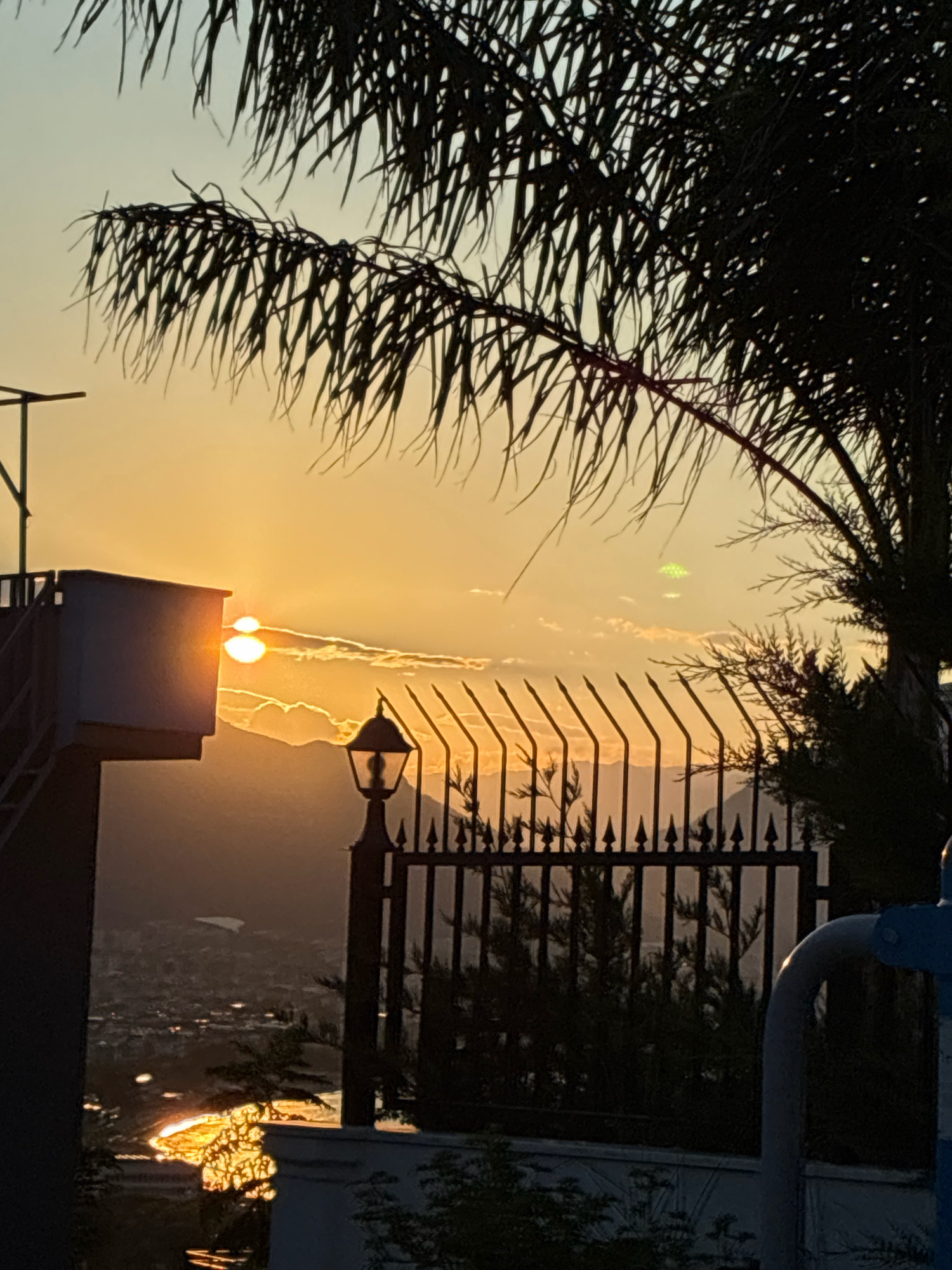 The image captures a serene sunset over Alanya, Turkey. The sky is painted with hues of orange and yellow, with soft clouds scattered across it. In the foreground, a palm tree's fronds are silhouetted against the vibrant sky. A dark metal fence with pointed finials runs across the middle ground, with a traditional-style lantern positioned just inside it. To the left, a white structure with a metal railing and stairs is visible, partially obscuring a view of the city below, which is dimly lit by the setting sun. The distant mountains are silhouetted against the horizon. The overall scene evokes a sense of tranquility and natural beauty.