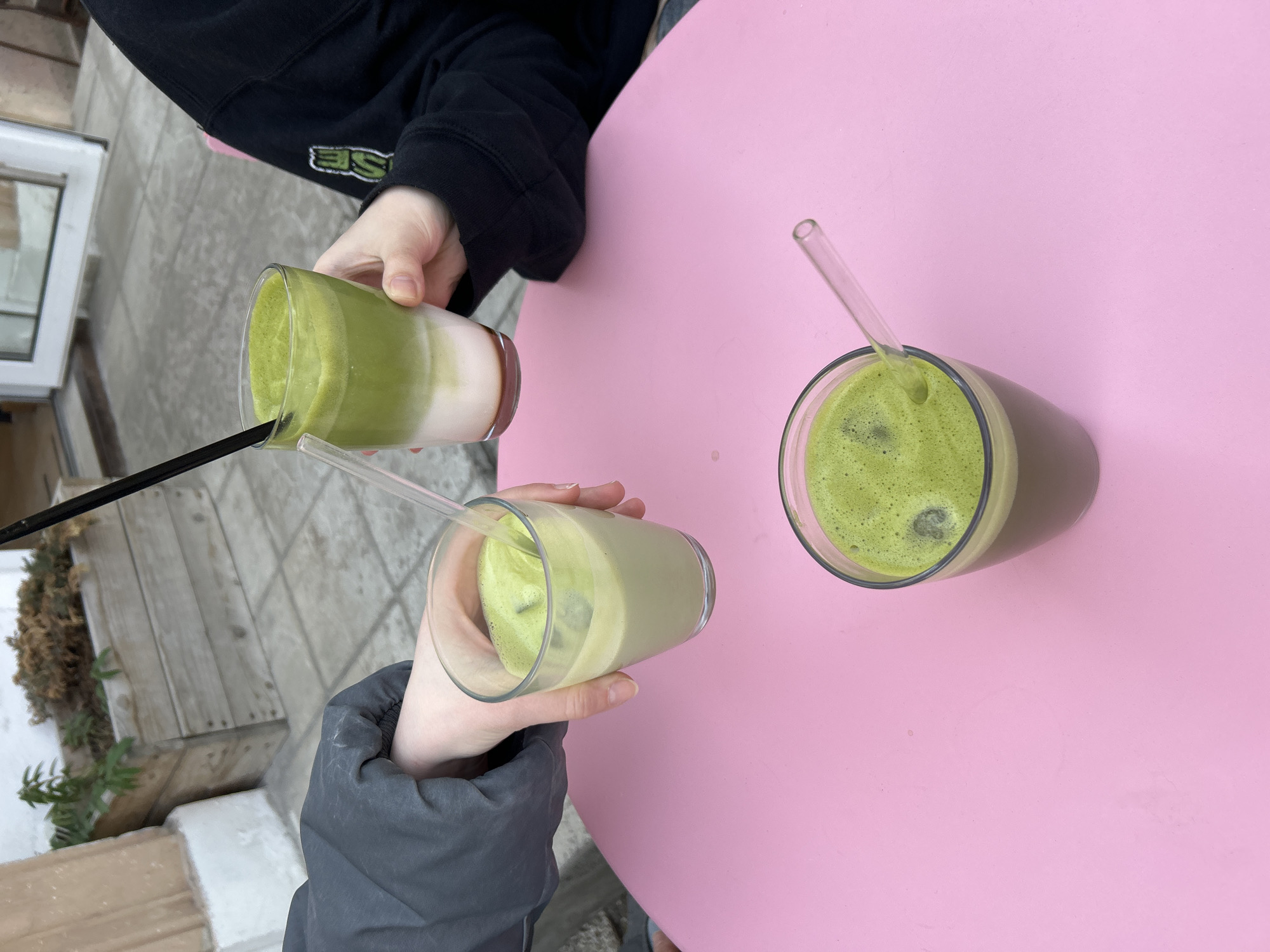 The image shows three drinks on a pink table. Two of the drinks are in clear glasses, one is being held by a person wearing a gray jacket, and the other is being held by a person wearing a black hoodie. Both drinks appear to be matcha lattes with ice and clear straws. The third drink is a matcha latte with no ice, placed on the table, with a clear straw. The location is an outdoor patio in Saint Petersburg, Russia. The weather is overcast.