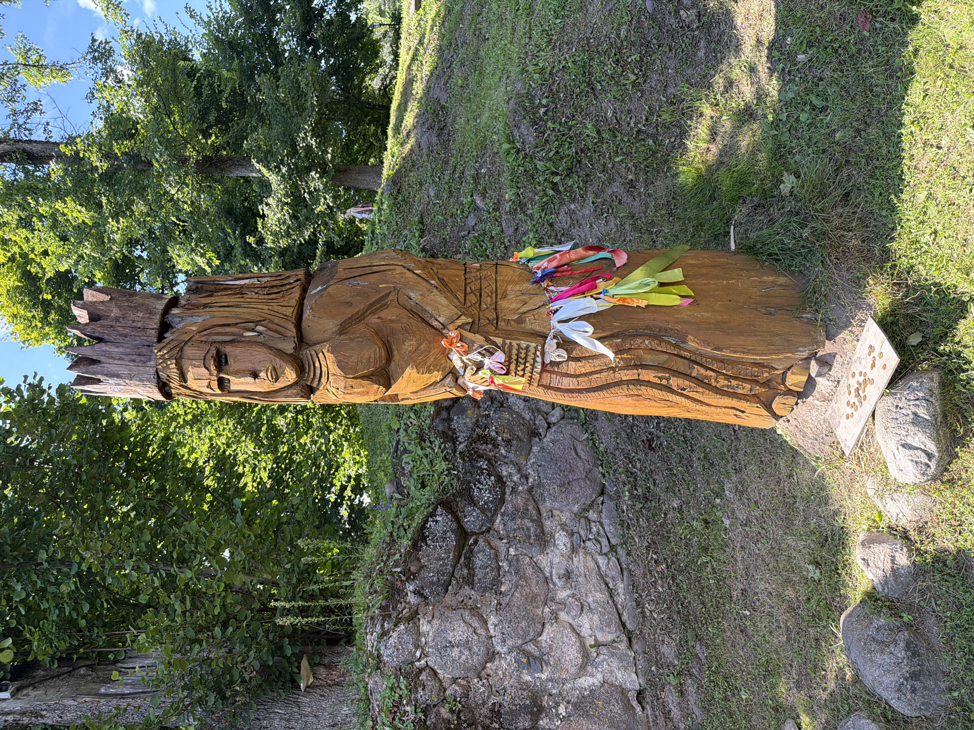 A carved wooden statue of a woman wearing a crown stands in a grassy area with trees in the background. The statue is brown and appears to be made of a single log. It has carved facial features, a torso, and a skirt. Colorful ribbons are tied around the statue's waist and a small woven basket is also attached there. In front of the statue, on a small wooden plank, are several coins. Behind the statue, a stone wall made of large, irregular stones is visible, with grass and small plants growing on and around it. The background consists of lush green trees and a patch of blue sky. A person is visible walking on a grassy hill in the distance. The overall scene suggests an outdoor setting in a park or natural area.