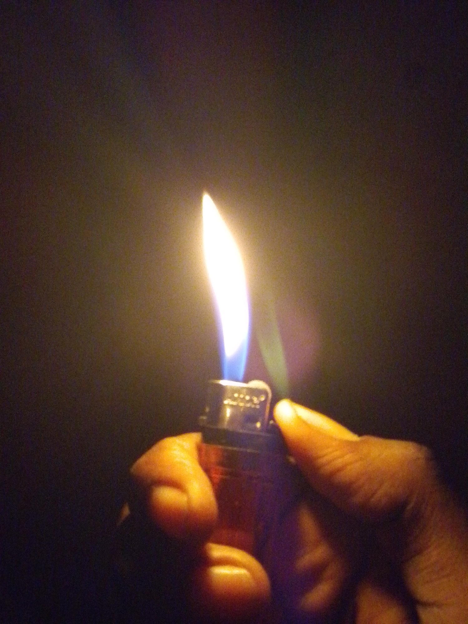 A close-up shot shows a hand holding a red lighter with a flame. The flame is bright orange at the base, fading to yellow and then white at the tip, with a hint of blue at the bottom. The background is dark and out of focus. The image appears to have been taken at night, in Serti, Nigeria.