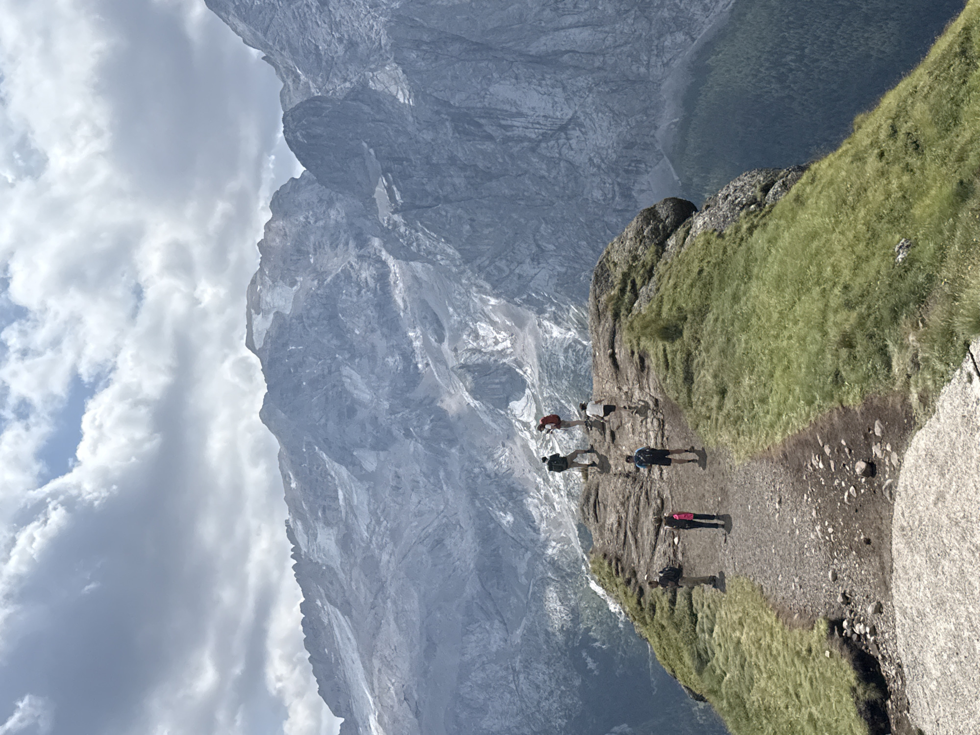 A vertical, wide-angle shot captures a group of seven hikers on a grassy, rocky mountain ridge with a steep drop on the right. In the background, a vast, rugged mountain range under a cloudy sky dominates the scene. The hikers, some with backpacks, are scattered across the ridge. Four are closer to the foreground, one wearing a bright pink backpack and another with a blue backpack. Further up the ridge, three more hikers are visible, one with a red backpack. The terrain transitions from a gravel path in the foreground to a rocky surface where the hikers stand, covered in green grass and patches of lighter-colored rock. The massive mountains in the background are grey and imposing, with areas of snow or glaciers on their peaks and steep, textured rock faces. The sky is filled with dramatic, grey and white clouds, with glimpses of blue sky peeking through. The overall impression is one of a challenging and scenic mountain hike. This scene is likely in the vicinity of the Marmolada glacier, which is near Canazei, Italy.