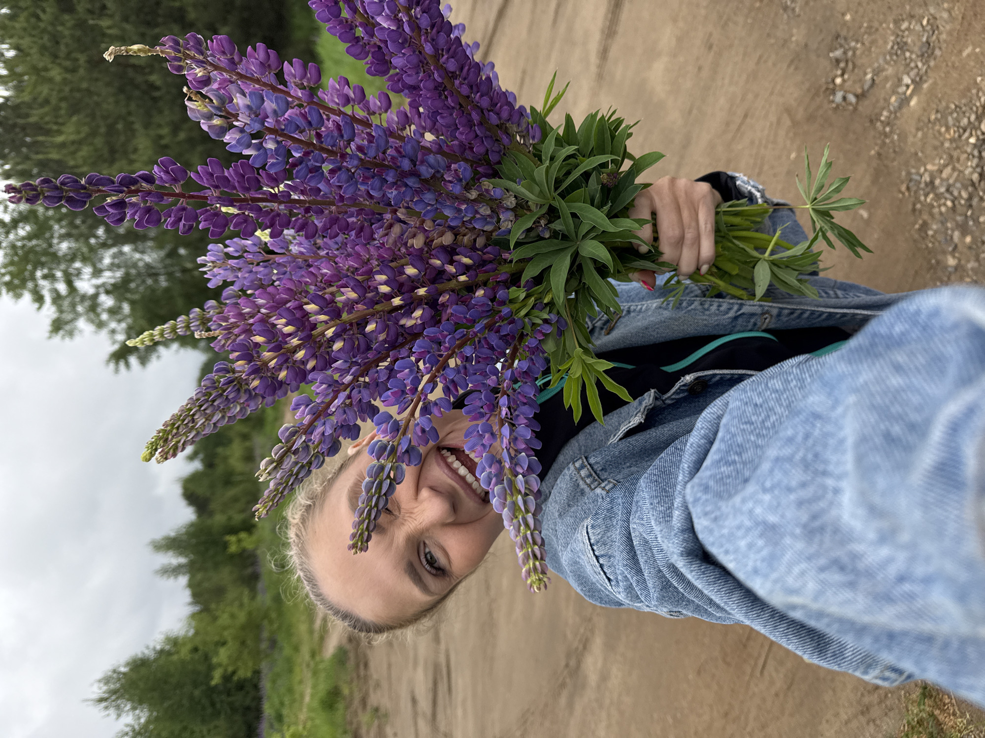 A woman is holding a bouquet of purple lupine flowers and smiling at the camera. She is wearing a denim jacket over a dark-colored shirt. The background shows a dirt road, greenery, and a cloudy sky. The image appears to be taken outdoors in a rural or natural setting. While a definitive determination of the location as Chaikovsky, Russia, is not possible, the environment is consistent with that general region.