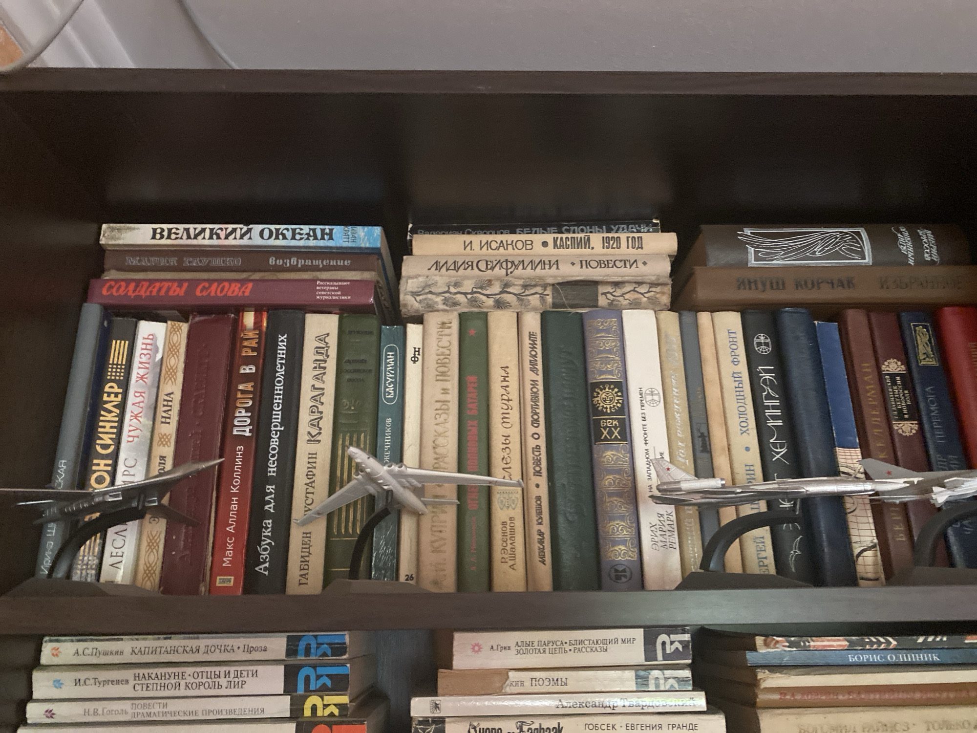 The image shows a dark brown wooden bookshelf filled with books and two model airplanes. The books are arranged in stacks and rows, with titles in Russian. On the top shelf, there are several hardcover books stacked horizontally. The middle shelf is filled with books standing upright, and two model airplanes are placed in front of them. The bottom shelf also contains books, with more model airplanes visible in the foreground. The lighting is dim, and the overall impression is of a cluttered bookshelf. The scene appears to be indoors, likely in a home or library.