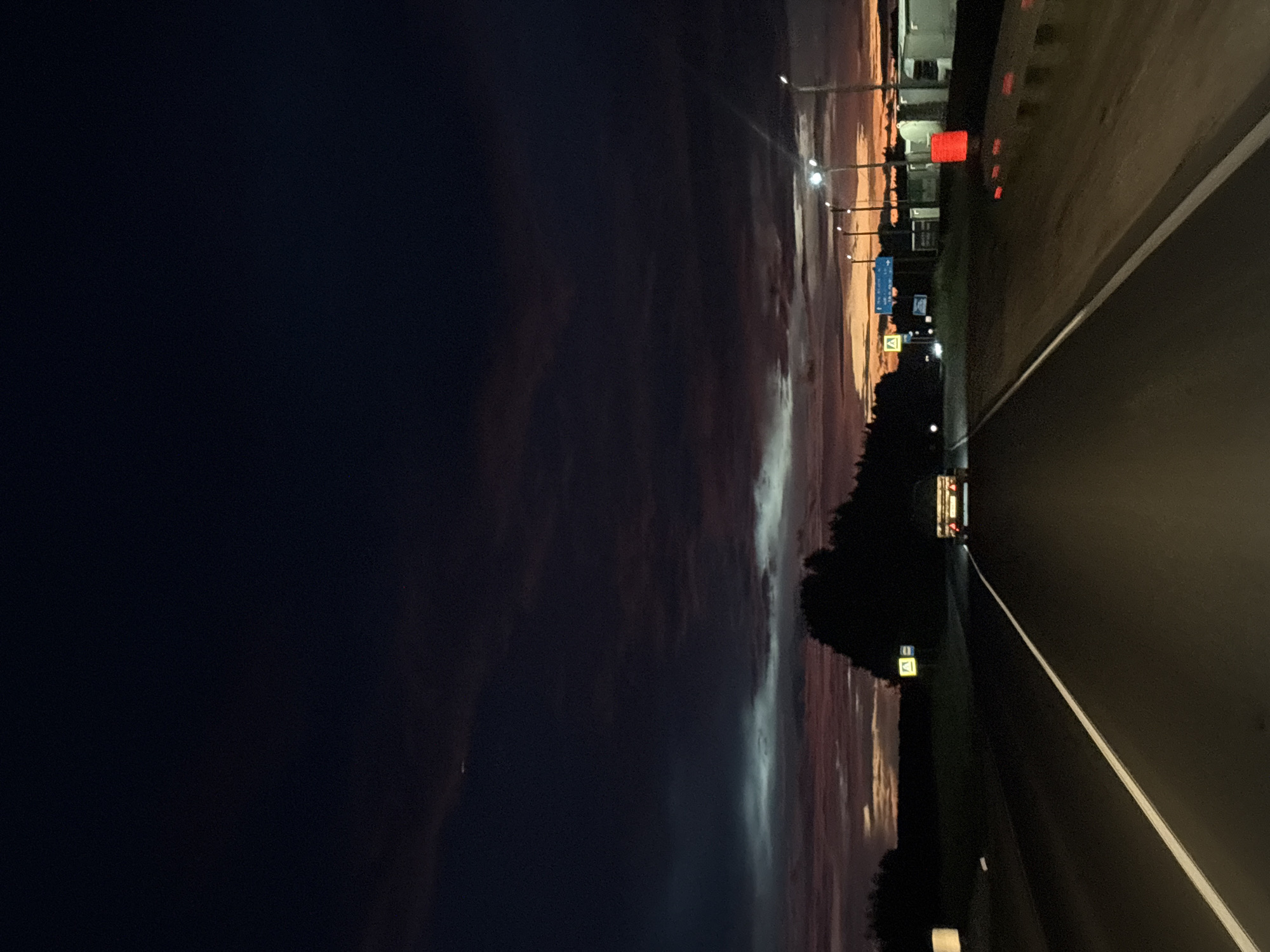 This media file depicts a nighttime scene on a highway in Kotelnich, Russia. The image is oriented vertically. The left side of the frame is dominated by a dark, almost black sky with some faint, wispy clouds that show subtle hues of purple and orange from the distant sunset or twilight. Streetlights are visible along the right side of the highway, casting pools of light.
The highway itself occupies the right side of the image, with a clear white dividing line. The road surface appears dark and wet, suggesting recent rain or dew. A vehicle, possibly a van or truck, is seen driving away in the distance on the highway, its taillights illuminated. Along the side of the road, there are various signs and structures. One prominent sign is blue with white text, and another is a yellow square sign with a black symbol inside. There are also red lights visible on what appears to be a barrier or structure along the roadside. Dark trees form a silhouette along the edge of the highway, further emphasizing the nighttime setting. The overall mood is one of a solitary journey at dusk or early night. The sky's colors suggest the time is either very late evening or very early morning.