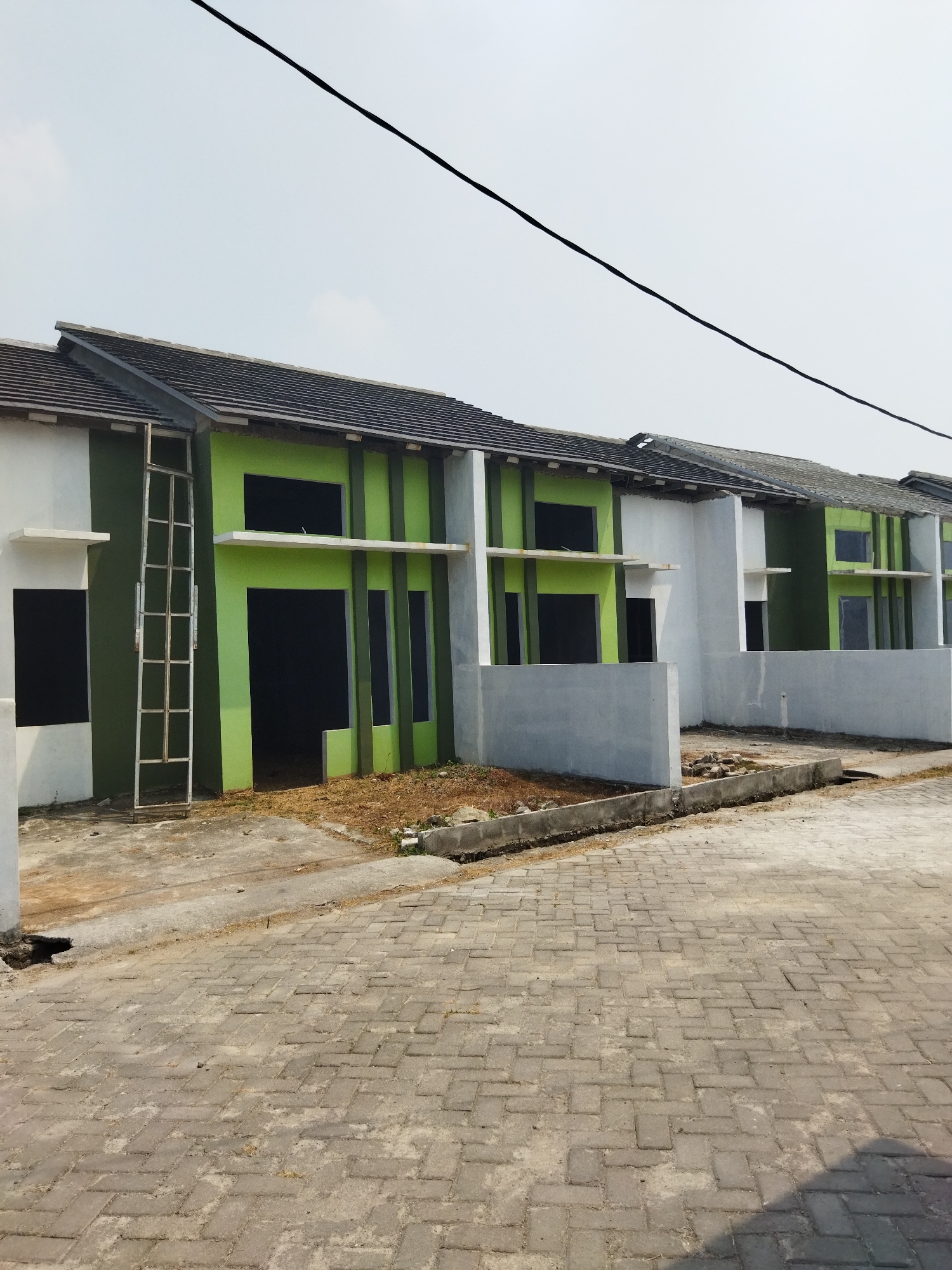 In the city of Suka Sari, Indonesia, a row of newly constructed houses sits along a paved street. The houses are a modern design, with white and bright green exteriors. Each house has a dark, tiled roof and dark rectangular windows. The street is paved with interlocking gray bricks. In the foreground, dry grass and dirt are visible. The sky is overcast and a thin black wire stretches across the top of the frame.