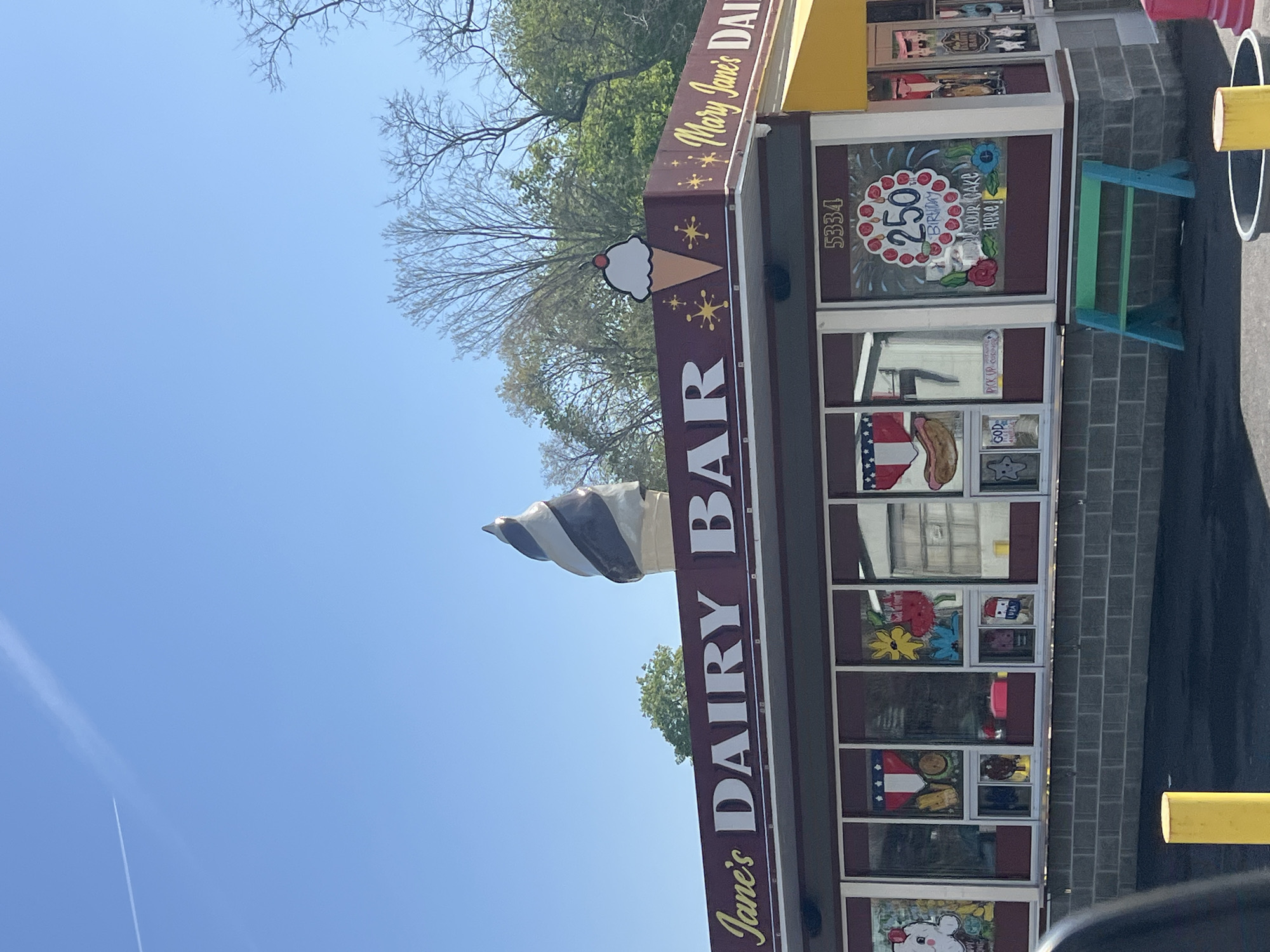 The image, rotated, depicts the vibrant exterior of Mary Jane's Dairy Bar under a clear blue sky. A large, realistic sculpture of a chocolate and vanilla swirl ice cream cone adorns the roof, alongside a smaller, flat ice cream cone sign topped with a cherry. The building's windows are extensively decorated with colorful paintings, including an American flag motif, a hot dog, various flowers, and cartoon figures. One prominent window features a large birthday cake design with "250 BIRTHDAY YOUR CAKE HERE" visible, indicating a special event or offer. The address "5334" is also displayed above a window. Other visible text includes "GOD" and "PICK UP C" on window decals. In front of the establishment, a small blue and green picnic bench is present, along with yellow bollards. The background shows bare tree branches, suggesting a seasonal transition, interspersed with some green foliage. A faint airplane contrail streaks across the bright sky. No people are visible, and the scene depicts a sunny day.