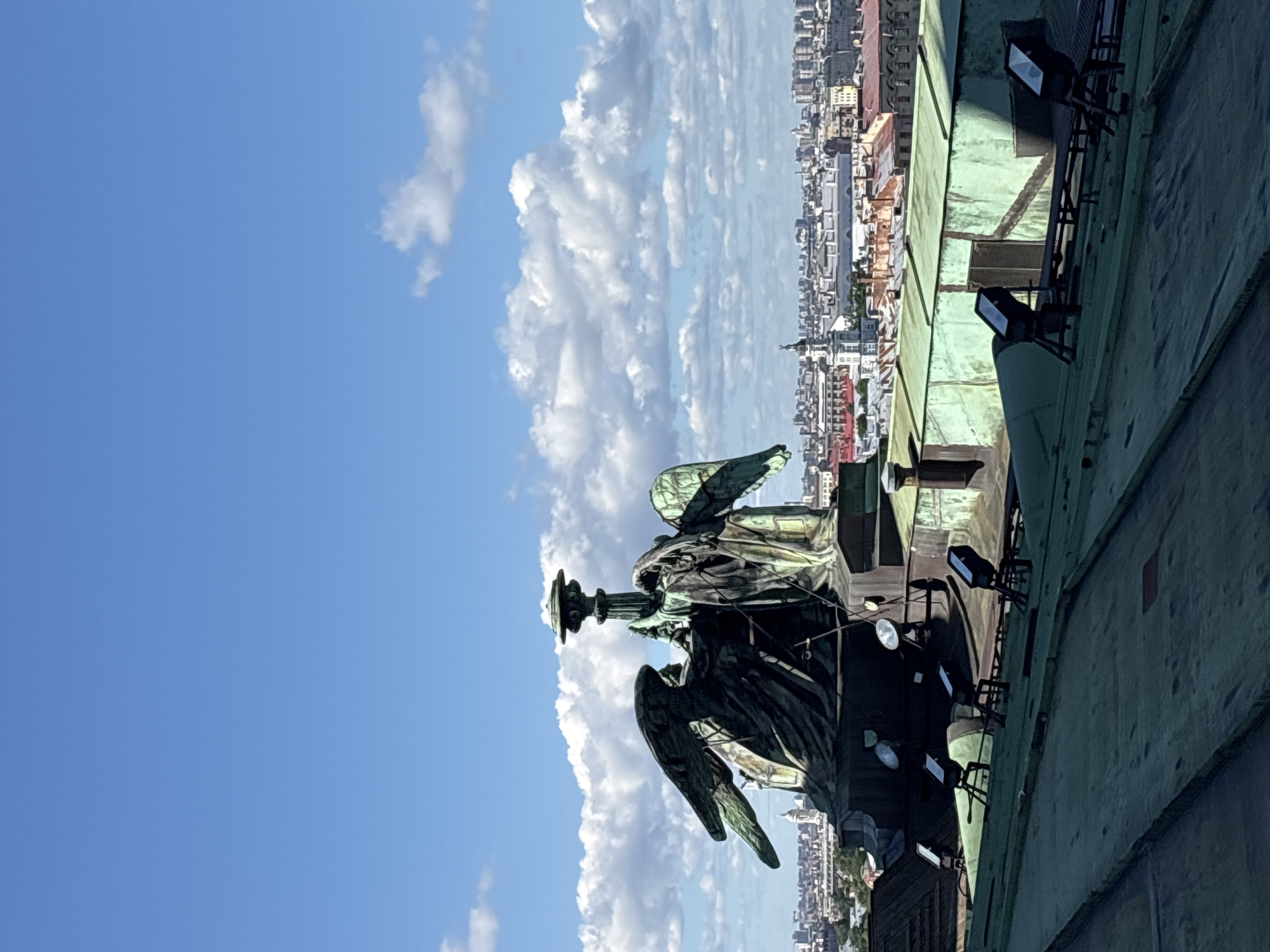 The image, captured vertically, shows a statue of an angel with outstretched wings, a common motif on the St. Isaac's Cathedral in Saint Petersburg, Russia. The angel, made of aged bronze, stands prominently in the foreground. Behind the statue, a panoramic view of the city unfolds under a bright blue sky dotted with fluffy white clouds, suggesting a clear, pleasant day. The city skyline is a dense tapestry of buildings with red and grey rooftops, characteristic of historical European architecture. In the immediate right of the frame, the weathered, green patina of a building's roof is visible, with several industrial-looking spotlights mounted on it, indicating the urban environment of a large city, likely taken from a high vantage point. The statue itself appears to be holding a staff or a torch. The lighting suggests it is daytime, possibly late morning or early afternoon given the brightness and the shadows cast. No visible text is present in the image.