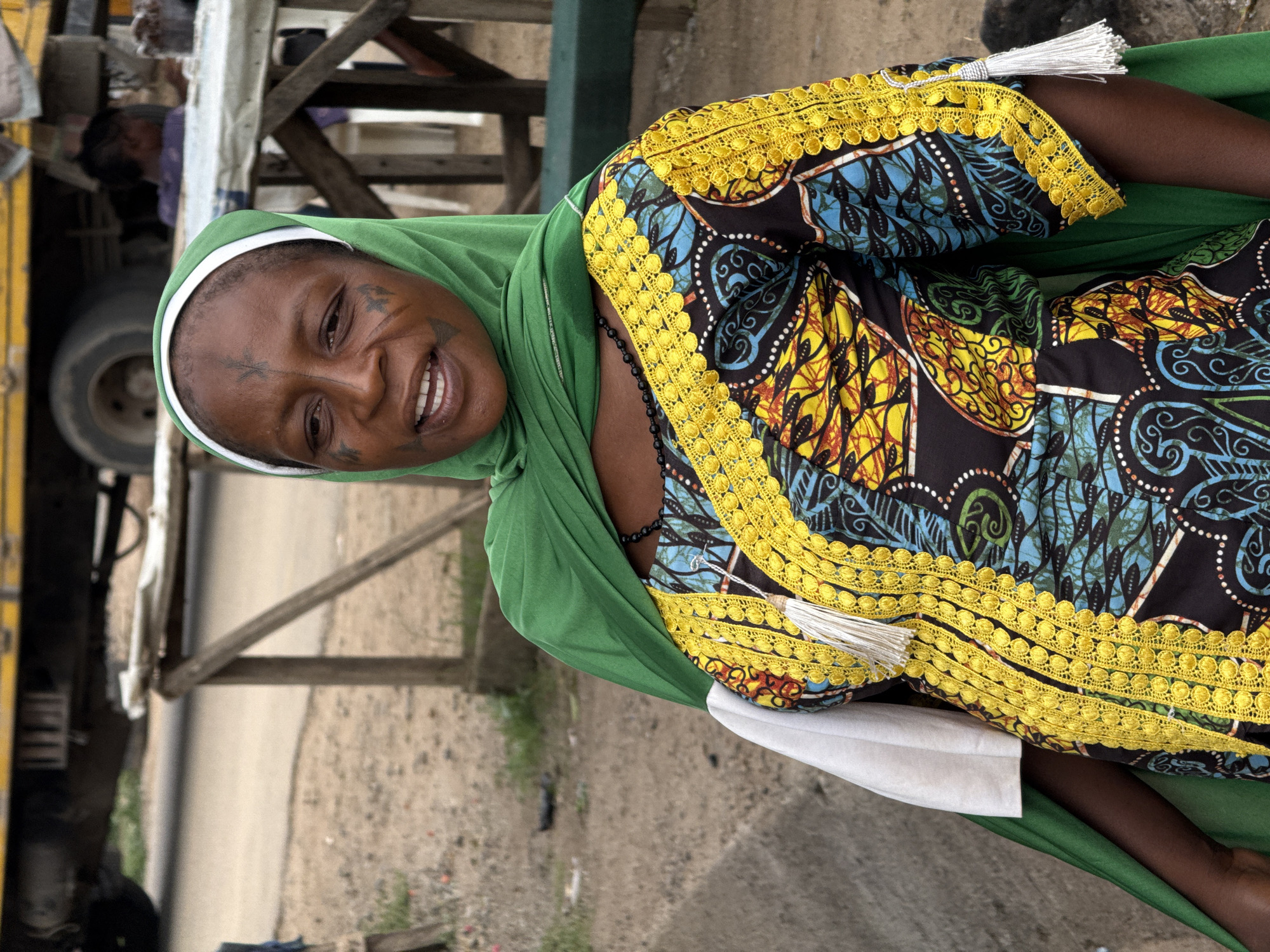 A medium shot, taken during daylight, shows a Black woman in Abuja, Nigeria. She is smiling directly at the camera. Her face has traditional tribal markings. She wears a bright green headscarf. Her dress is patterned and has a yellow lace trim. White tassels are attached to the sleeves. She has a black beaded necklace. A partially visible yellow and silver vehicle is in the background. The ground and various structures indicate an urban setting.