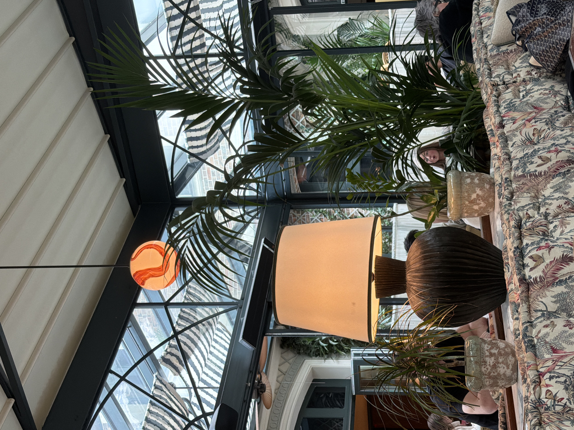 The image shows a well-lit interior scene, likely a restaurant or lounge in Paris, France. The primary subjects include a large lamp with a beige shade and a dark, ridged base, along with numerous potted plants, primarily palms. A patterned sofa is visible in the foreground. The architecture includes a glass ceiling with metal supports, suggesting an atrium or conservatory. Outside the glass ceiling, awnings are visible. People can be seen, including faces that appear to be patrons and staff of the establishment. The environment suggests an upscale setting. An orange, globe-shaped pendant light hangs from the ceiling.