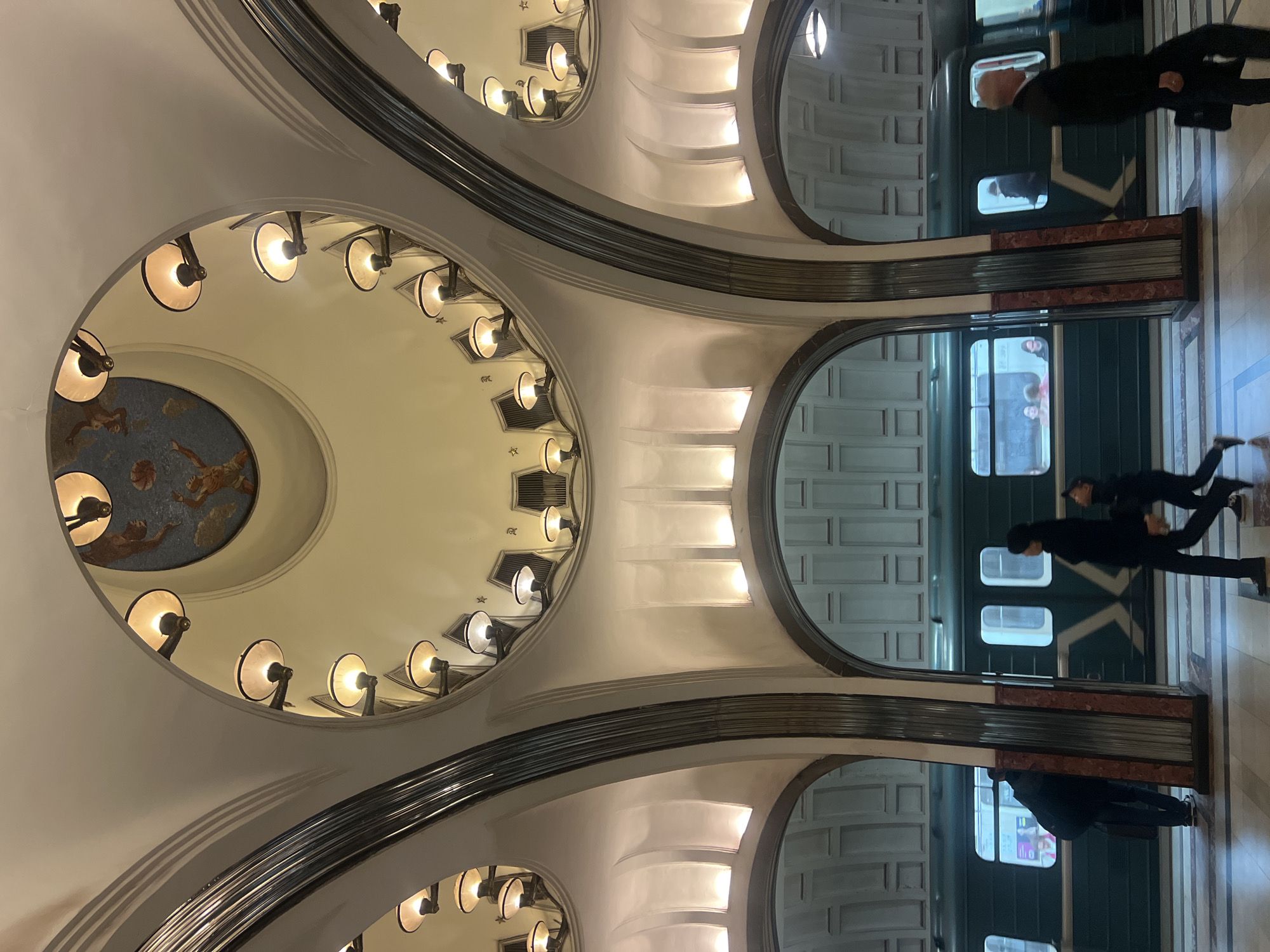 The image shows a subway station in an unknown city. The architecture is characterized by curved ceilings adorned with circular patterns of lights, and a round mosaic in one section. Two subway trains are visible along a platform, with people walking near them. The platform is marked with marble pillars and white walls with rectangular panels.
