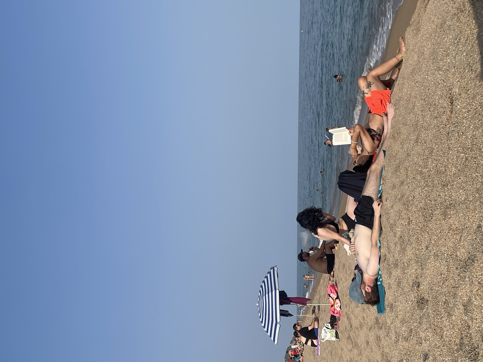 The image depicts a sunny day on the beach of Canet De Mar, Spain. The foreground shows two men and a woman relaxing on the sand. One man, wearing a blue baseball cap and black swim trunks, lies on a turquoise towel. Beside him, another man in black swim trunks is seated, with a woman in a black bikini next to him. The woman is looking away from the camera. Further up the beach, a striped umbrella provides shade for a man and woman sitting on the sand. In the background, the calm blue sea stretches to the horizon under a clear sky, with several people swimming and wading in the water.