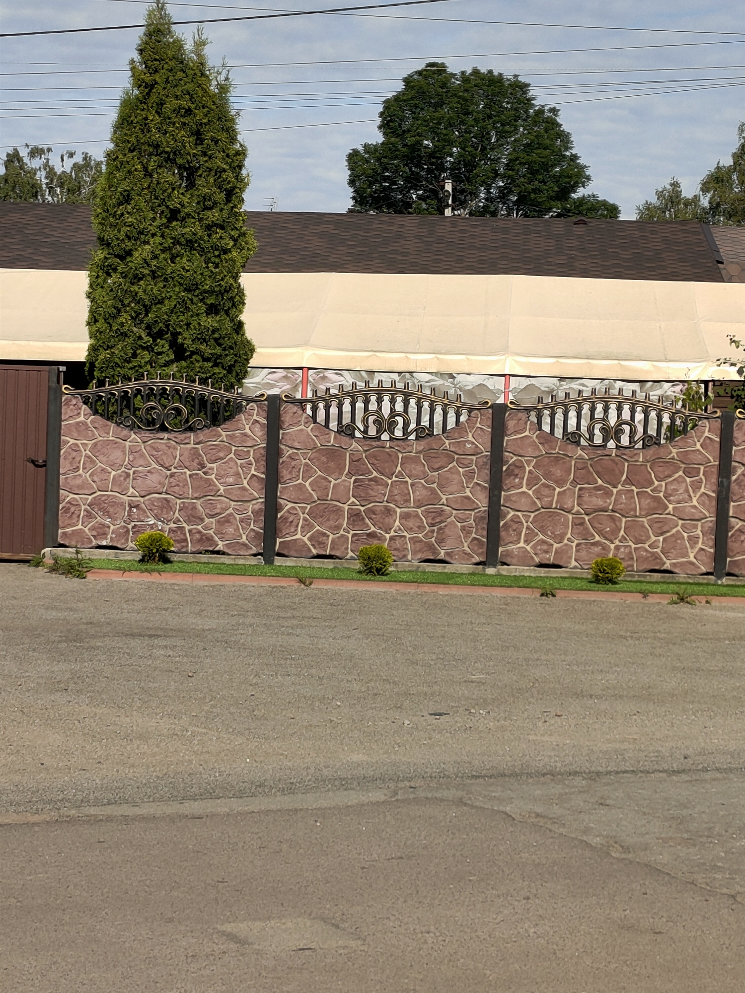 A tall, conical evergreen tree stands to the left of a decorative stone fence with ornate black and gold metalwork at the top. The fence is constructed of large, irregular reddish-brown stones with lighter grout lines, supported by dark vertical posts. Small, manicured green bushes are planted in front of the fence. Behind the fence, a building with a brown shingled roof is partially visible, covered by a light-colored awning or canopy. The ground in front of the fence is a strip of green grass and a paved area, which transitions into a dark asphalt road in the foreground. The sky above is overcast with scattered power lines. This scene is located in the city of Nosivka, Ukraine.