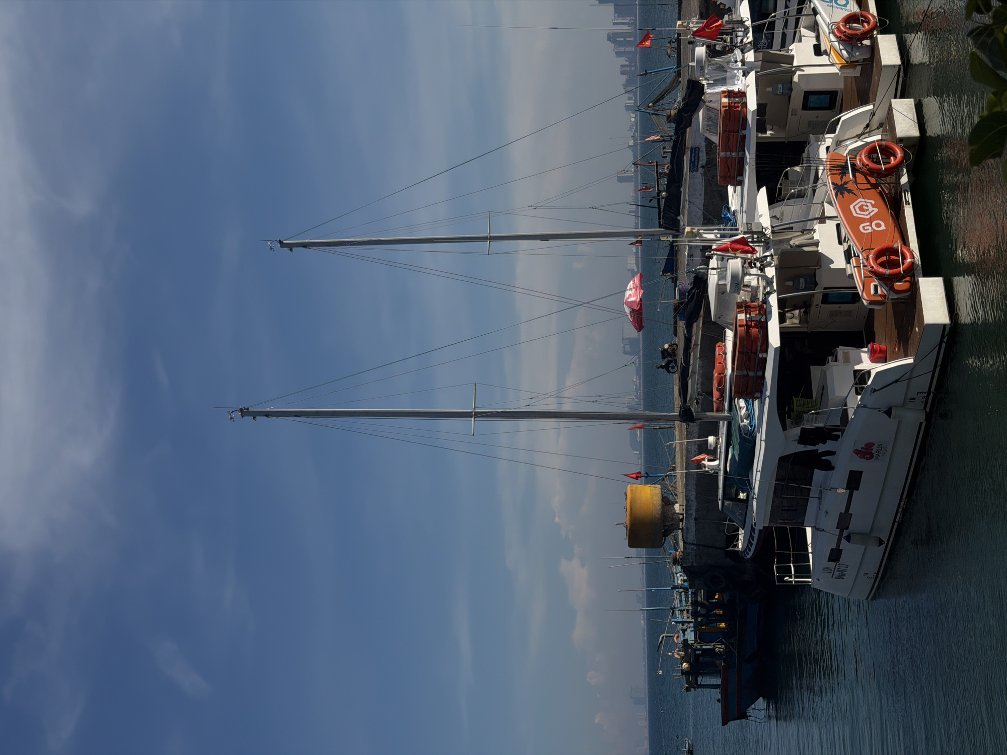 The image shows a marina with several boats docked, likely in Đà Nẵng, Vietnam, given the presence of Vietnamese flags and the city skyline visible in the background. A few of the boats are white catamarans, with safety equipment like life preservers and life rafts visible on their decks. A paddleboard with the letters "GQ" is attached to one boat. Other boats are smaller, with blue hulls. The sky is blue with wispy clouds. Two masts are visible, likely belonging to sailboats docked along the marina. The boats are moored next to a concrete pier. Some foliage is seen to the upper right.