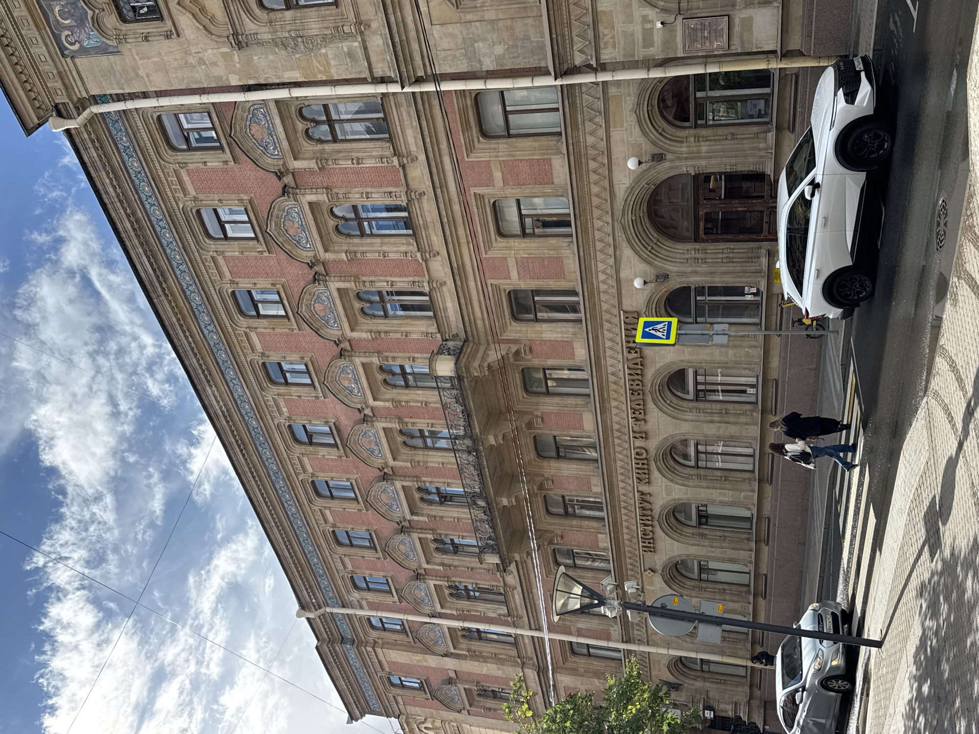 This is a street-level, eye-level, medium shot of a historic building in Saint Petersburg, Russia. The building, made of brick with stone accents, has a pink facade adorned with intricate blue and gold tilework along the upper levels and decorative carvings around the windows. The building's inscription in Russian reads "ИНСТИТУТ КИНО И ТЕЛЕВИДЕНИЯ," indicating it is the Institute of Film and Television.
In the foreground, a pedestrian crosswalk is visible, with two women walking across it from right to left. To the left of the crosswalk, a silver sedan is stopped at the intersection. A white SUV is parked further down the street on the right. A street lamp stands prominently on the left side of the image, casting a shadow on the pedestrian walkway. The sky above is blue with scattered white clouds and overhead electrical wires are present. A green tree is visible on the far left of the image. The street is paved with cobblestones and asphalt, with some wet patches suggesting recent rain. The overall scene depicts a typical urban streetscape in Saint Petersburg.